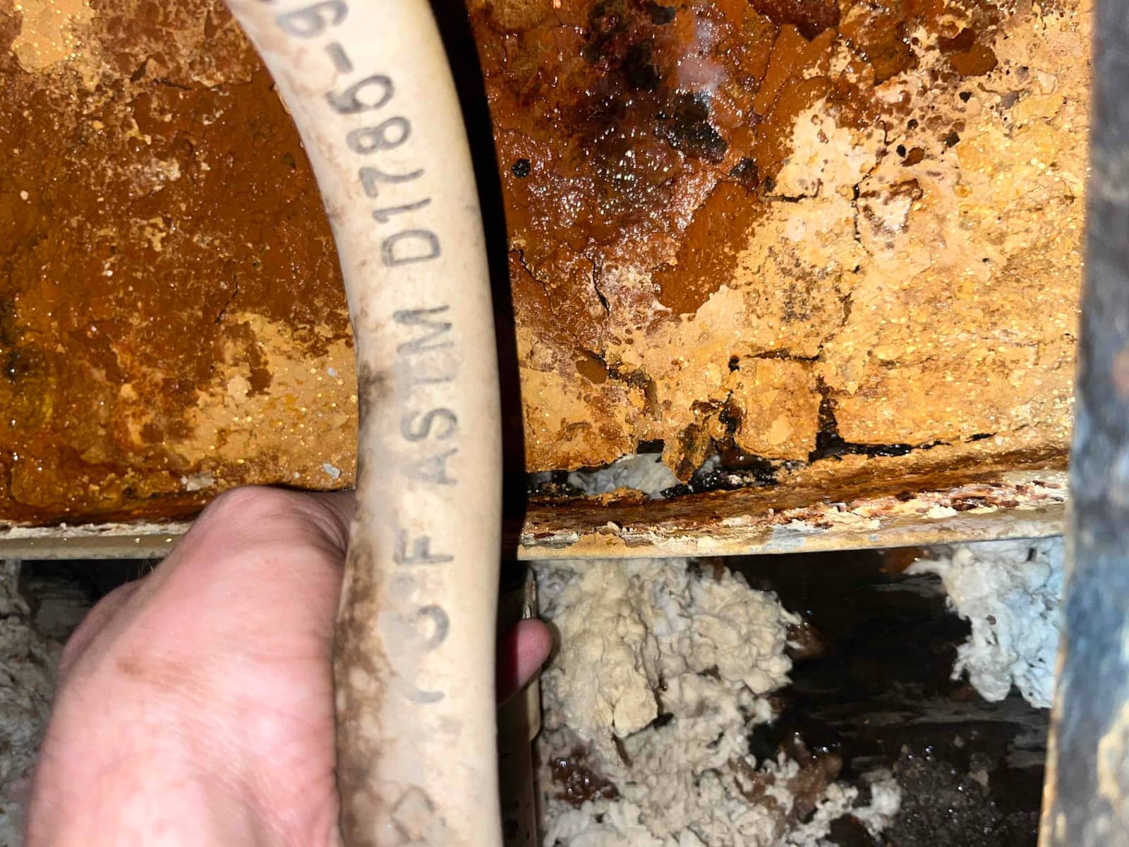 Corroded condensate drain pan with severe rust and mineral buildup, technician’s hand holding attached drain hose during replacement.