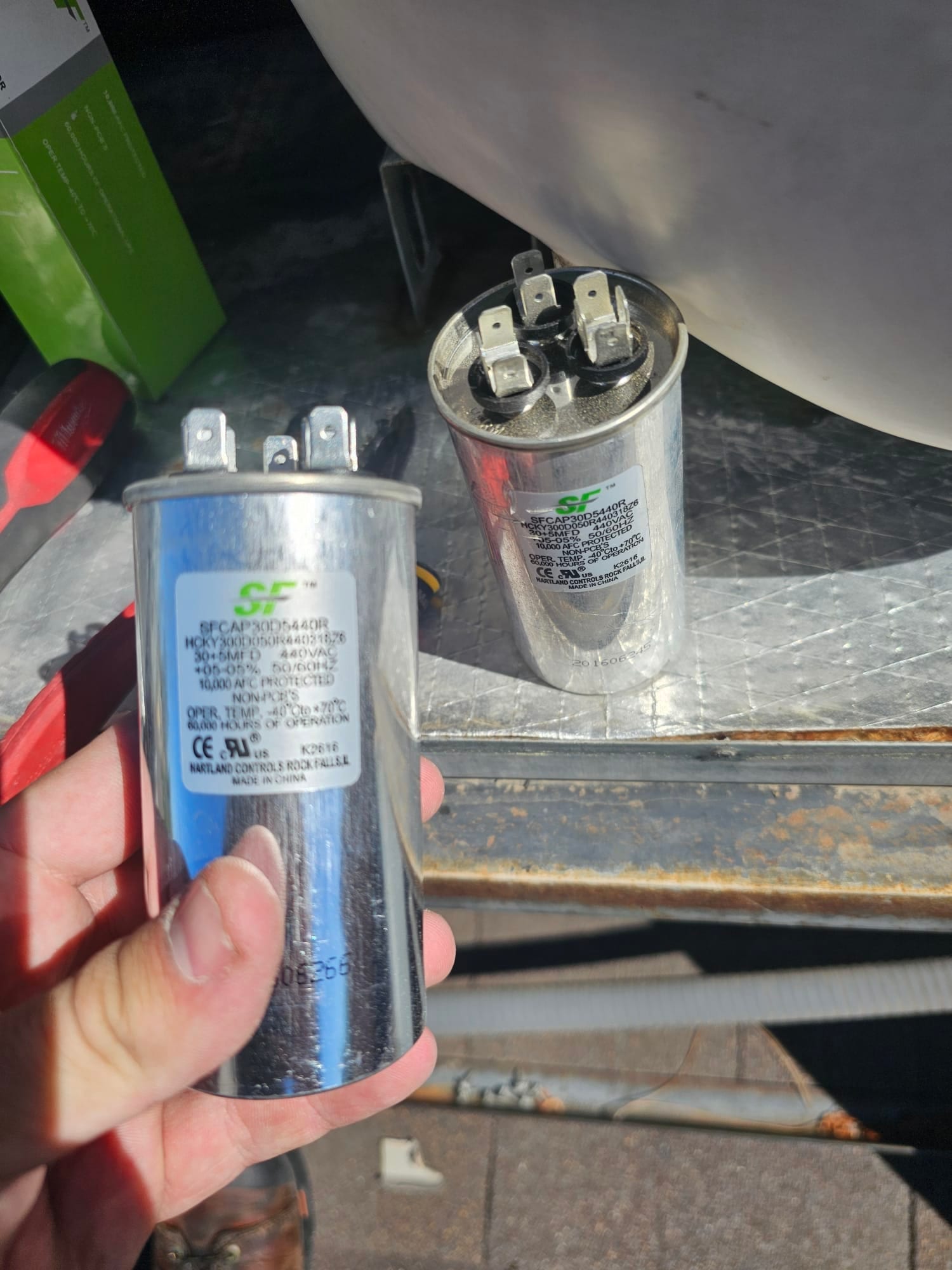 Two dual-run capacitors shown on the job site, each with three terminals and printed ratings visible on the labels.