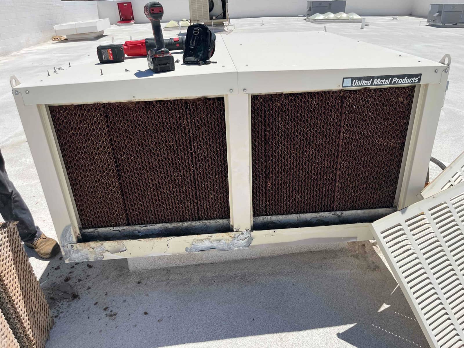 Evaporative cooler with old cellulose media pads removed, showing corroded lower trough and replacement pads staged beside the unit.