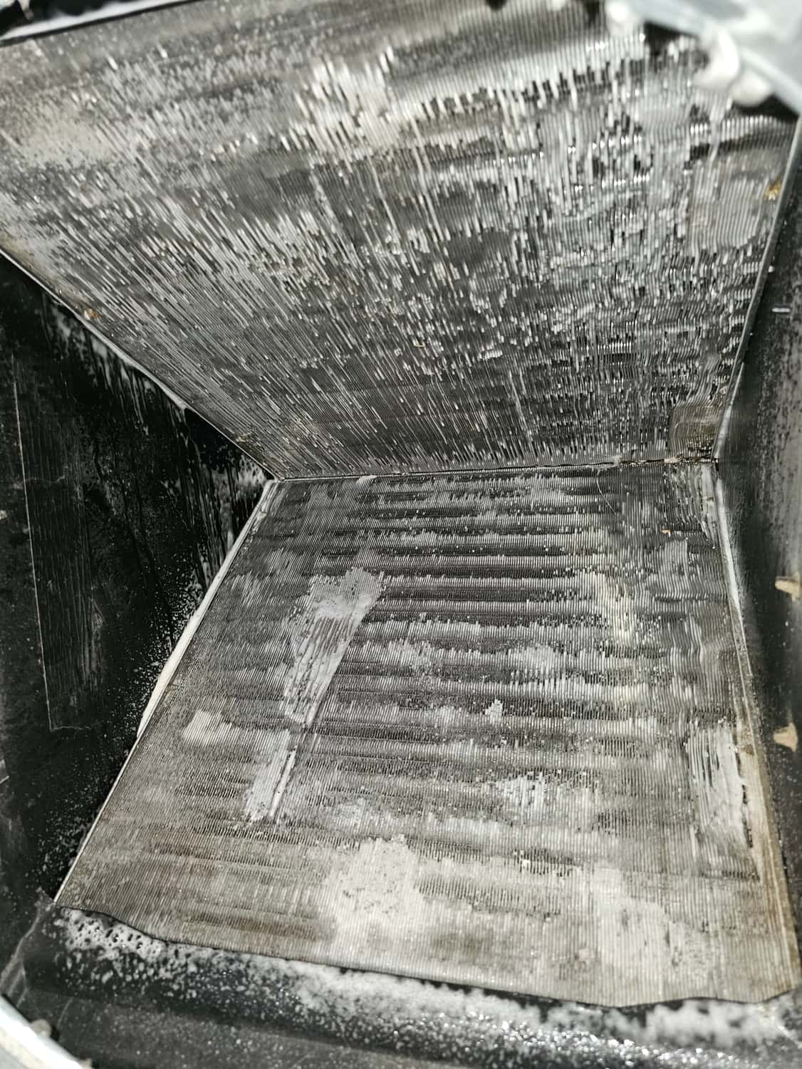 Indoor evaporator A-coil with aluminum fins visible, wet with foaming cleaner and moisture inside the drain pan area.