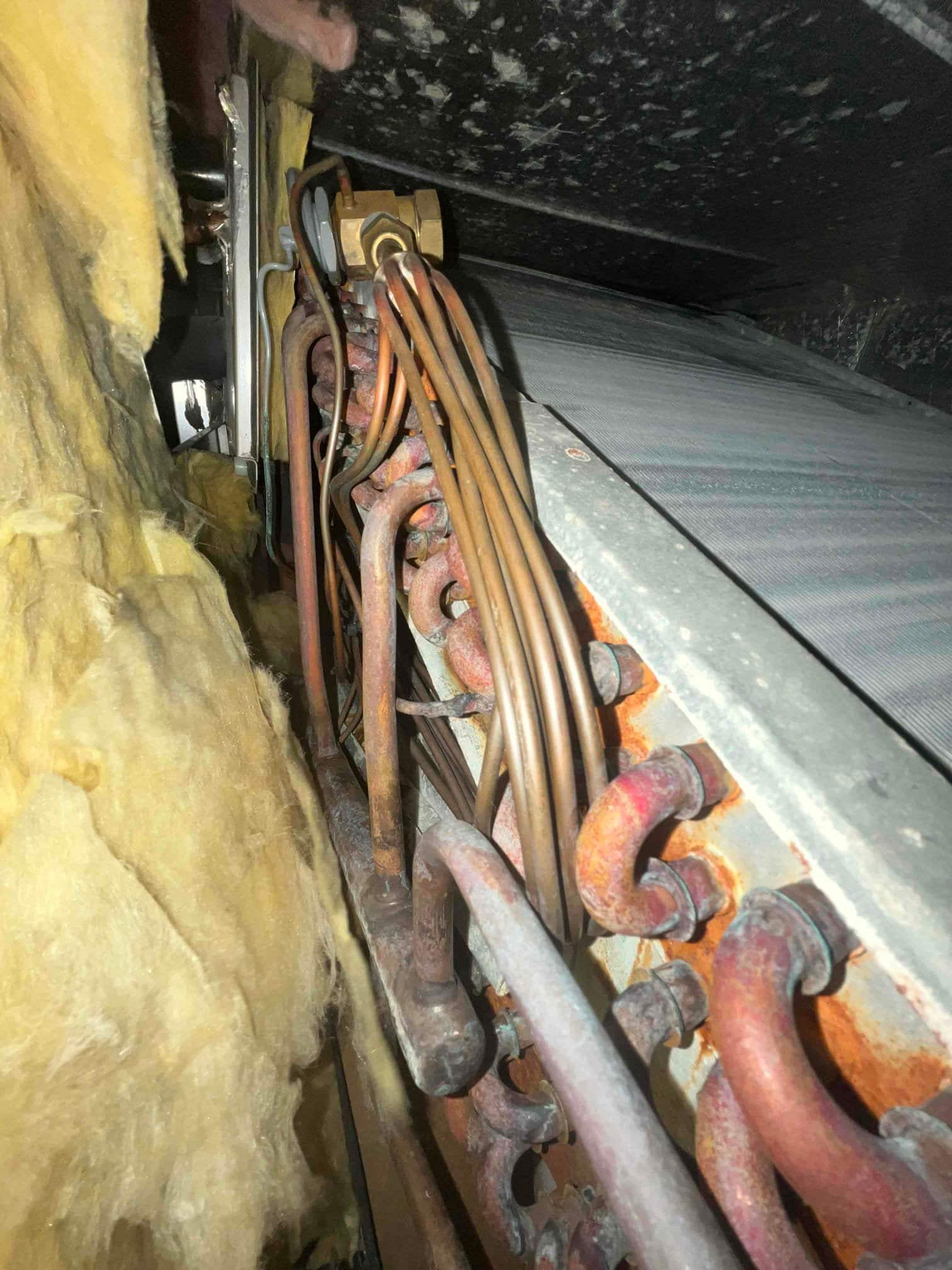 Indoor evaporator coil assembly with multiple copper refrigerant lines and rusted fittings, adjacent to exposed yellow fiberglass insulation.
