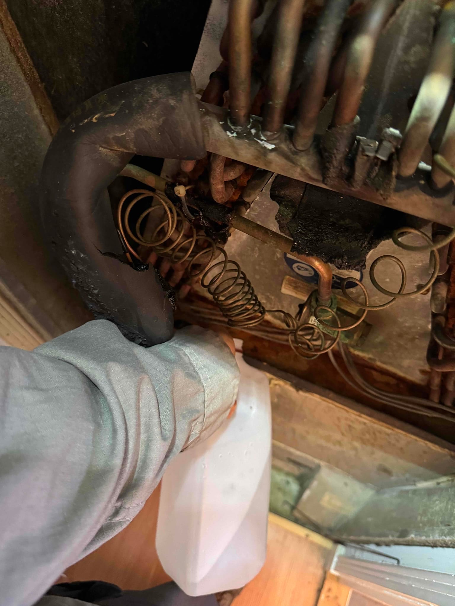 Indoor evaporator coil and condensate drain area with technician clearing water using a plastic container beneath rusted components.