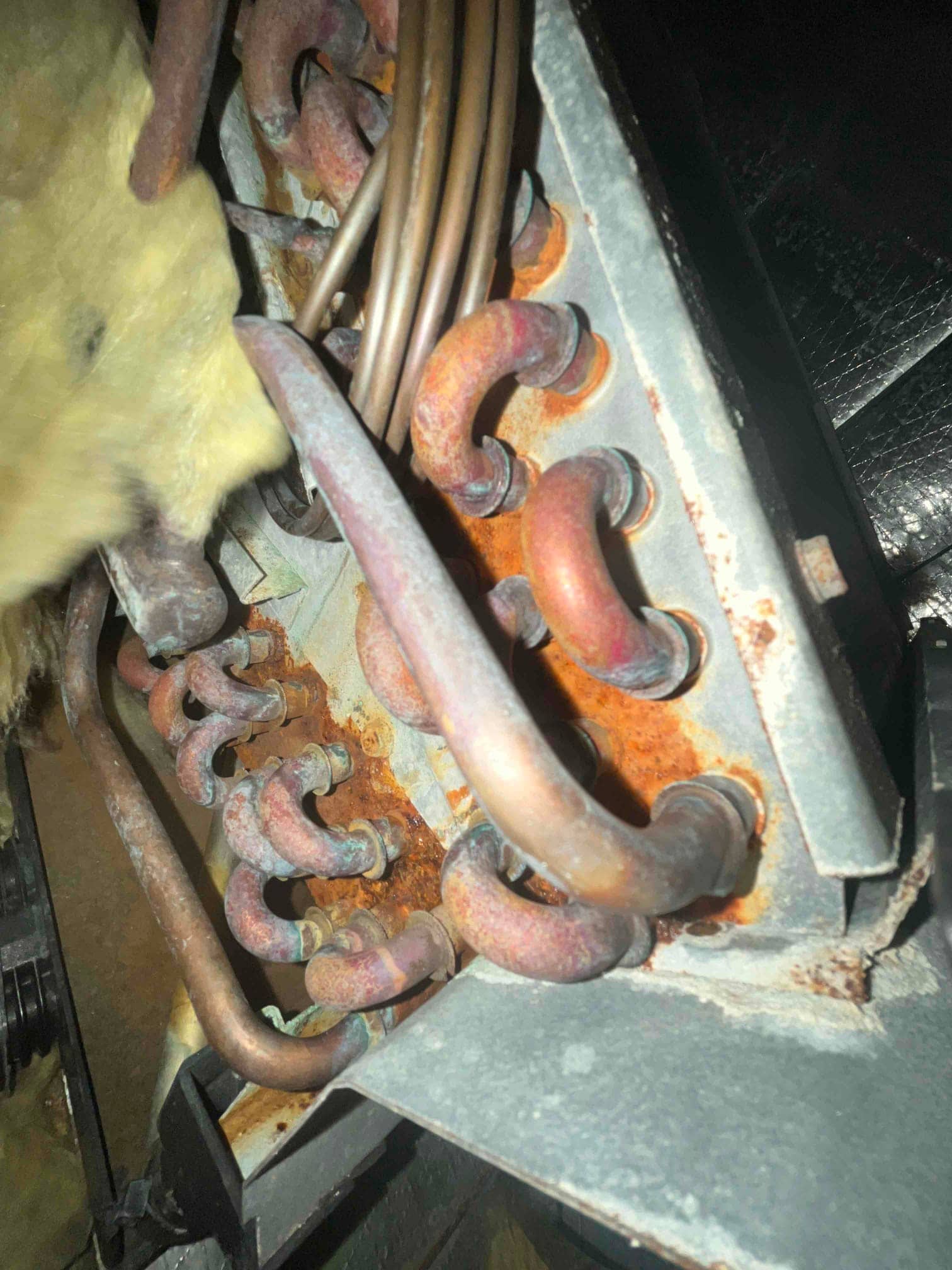Indoor evaporator coil tubing shows heavy rust and corrosion on multiple U-bends and metal surfaces near the plenum.