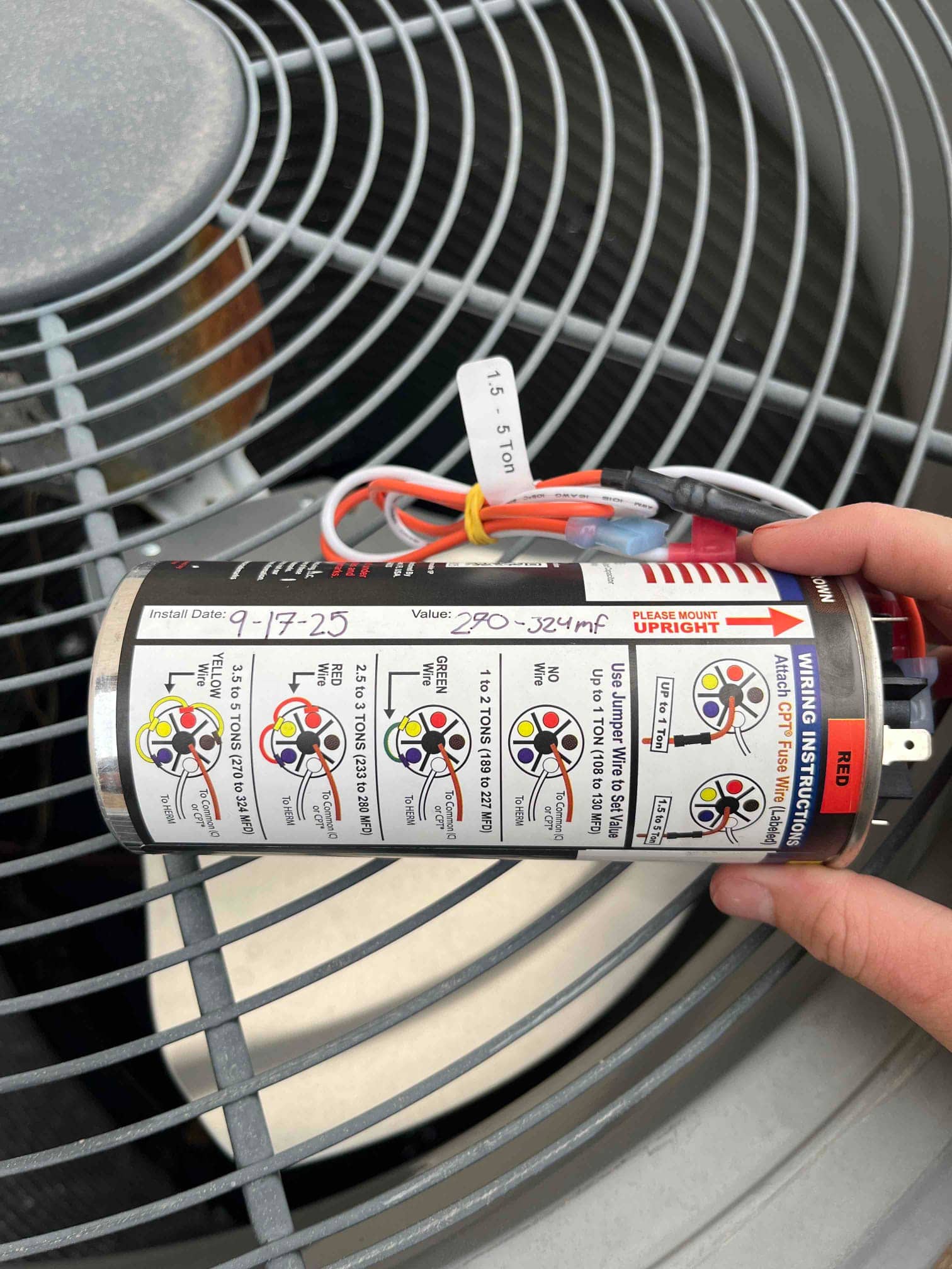New hard-start capacitor kit held beside the outdoor condenser fan, showing multiple wiring diagrams and labeled leads.