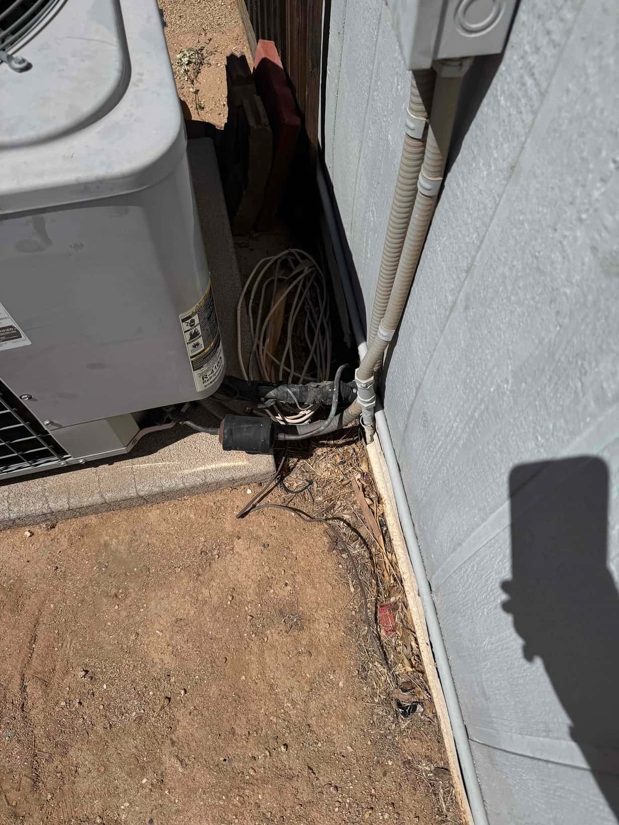Outdoor condenser next to a wall with exposed low-voltage wiring and a weatherproof disconnect box above the conduit.