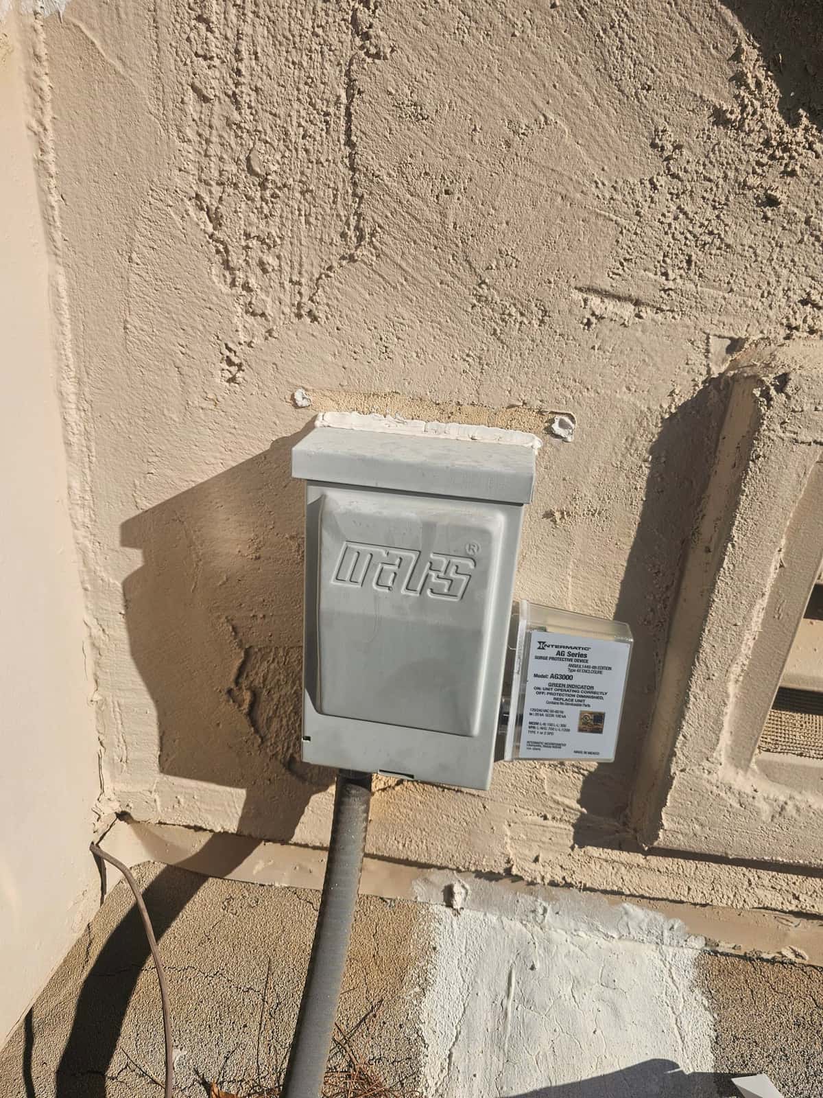 Outdoor HVAC electrical disconnect box with conduit entering from below, mounted on stucco wall near condenser location.