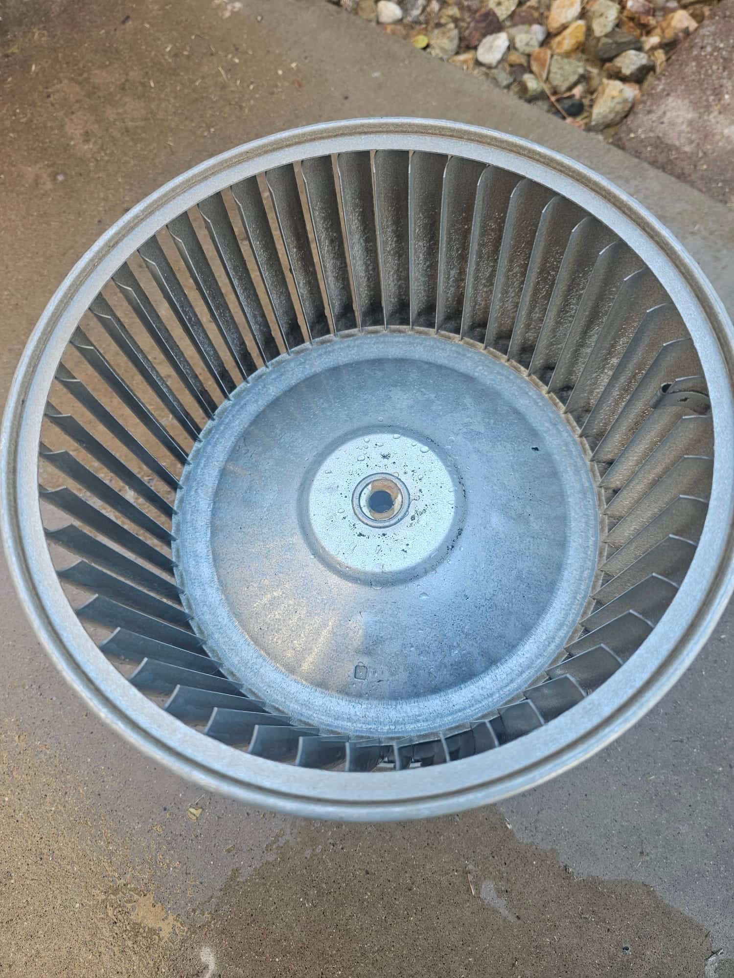 Removed blower wheel sits on the ground after cleaning, showing clean metal fins and hub with no debris.