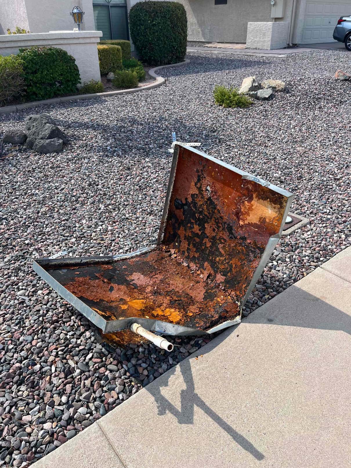Removed condensate drain pan sits on the ground, showing heavy rust, corrosion, and a connected drain outlet.