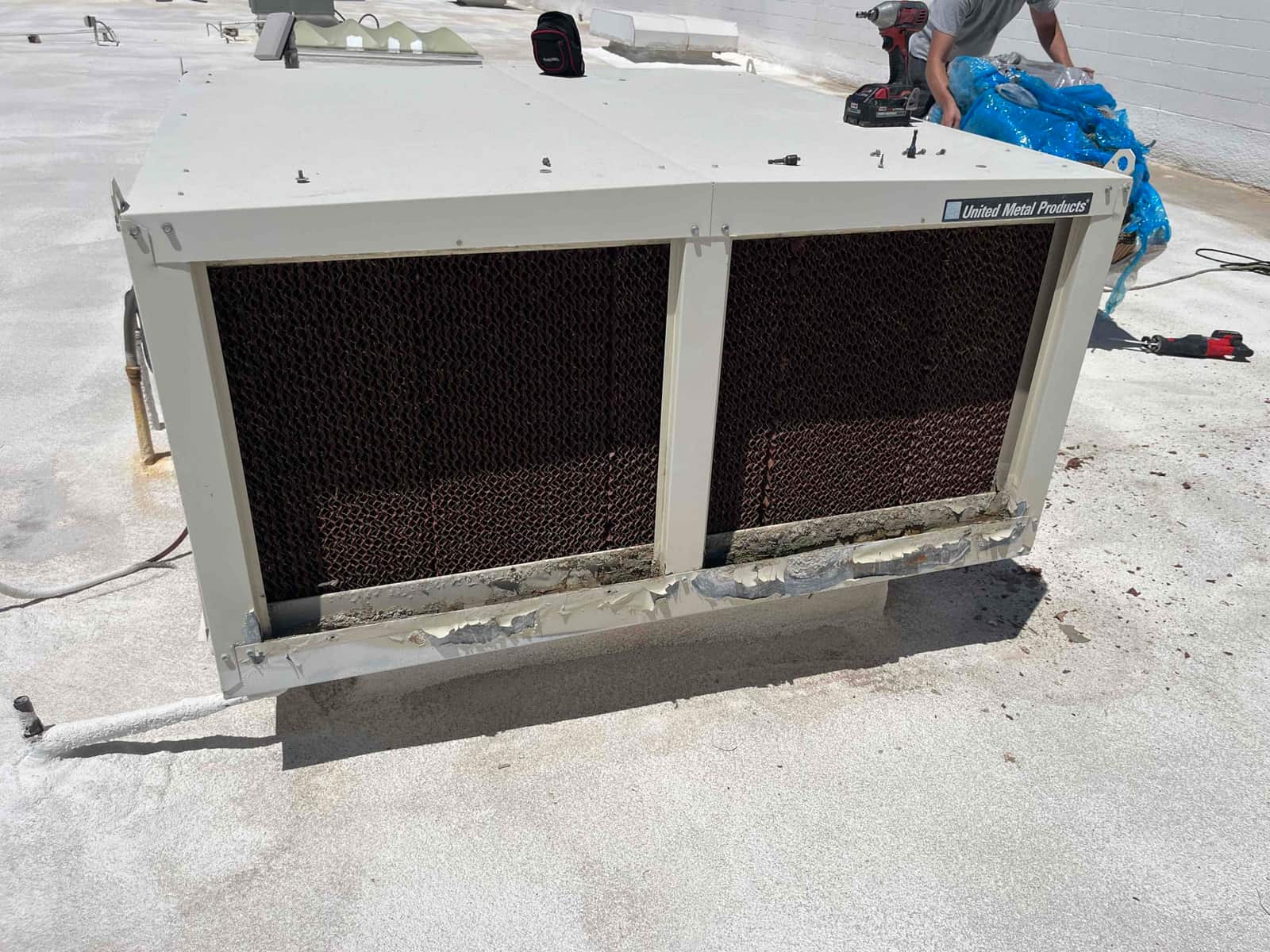 Rooftop evaporative cooler shows worn and corroded lower panel with exposed, aged media pads in the intake sections.
