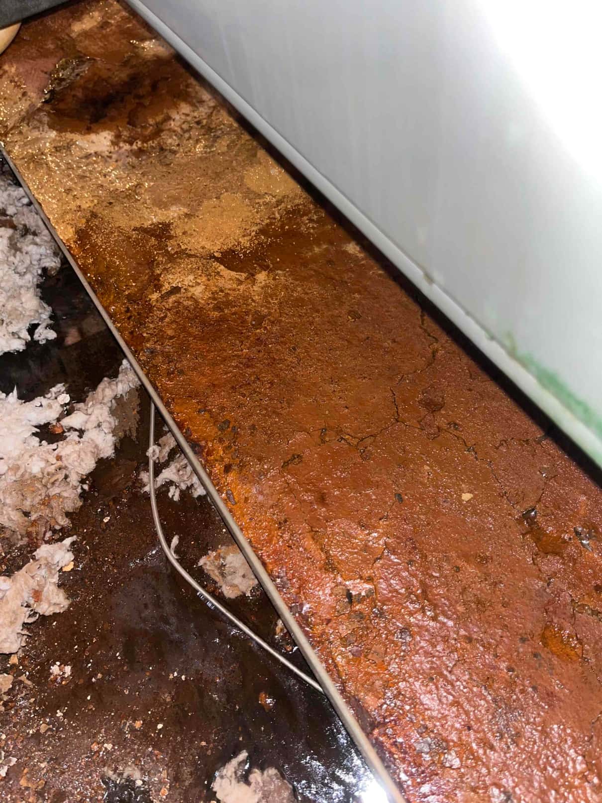 Rusted condensate drain pan under an air handler, showing heavy corrosion, standing water, and mineral deposits around the drain area.