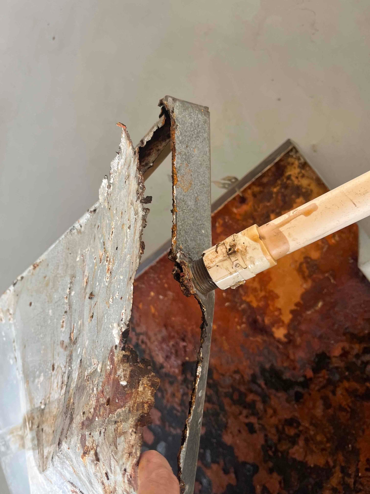 Rusted metal condensate drain pan removed, showing severe corrosion around the drain connection and deteriorated edges.