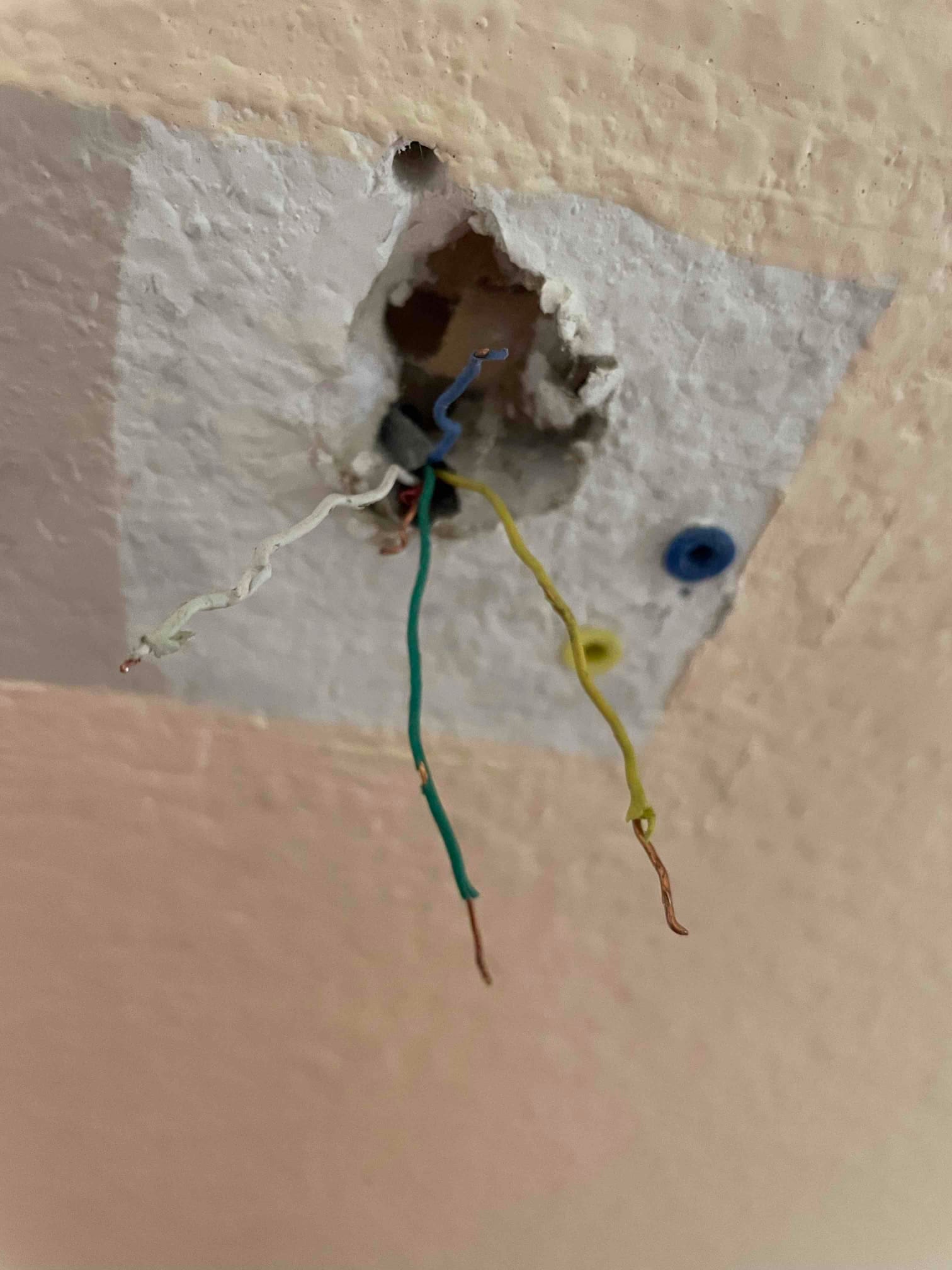 Stripped low-voltage thermostat wires—green, yellow, white, and blue—extend from a rough wall opening near a mounting patch.