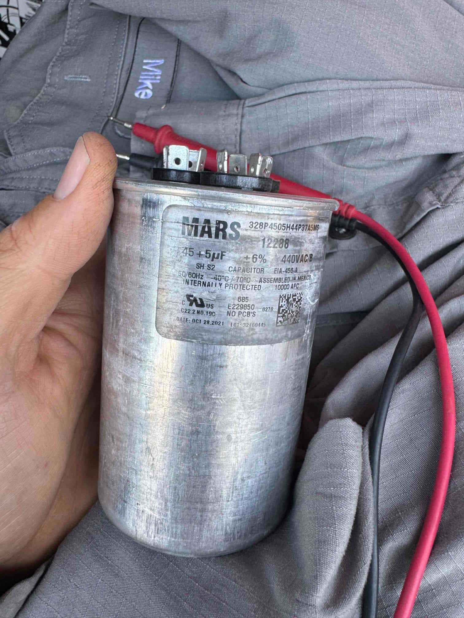 Technician holds a dual-run capacitor with clear terminals and printed rating label showing 45+5 μF, 440VAC.