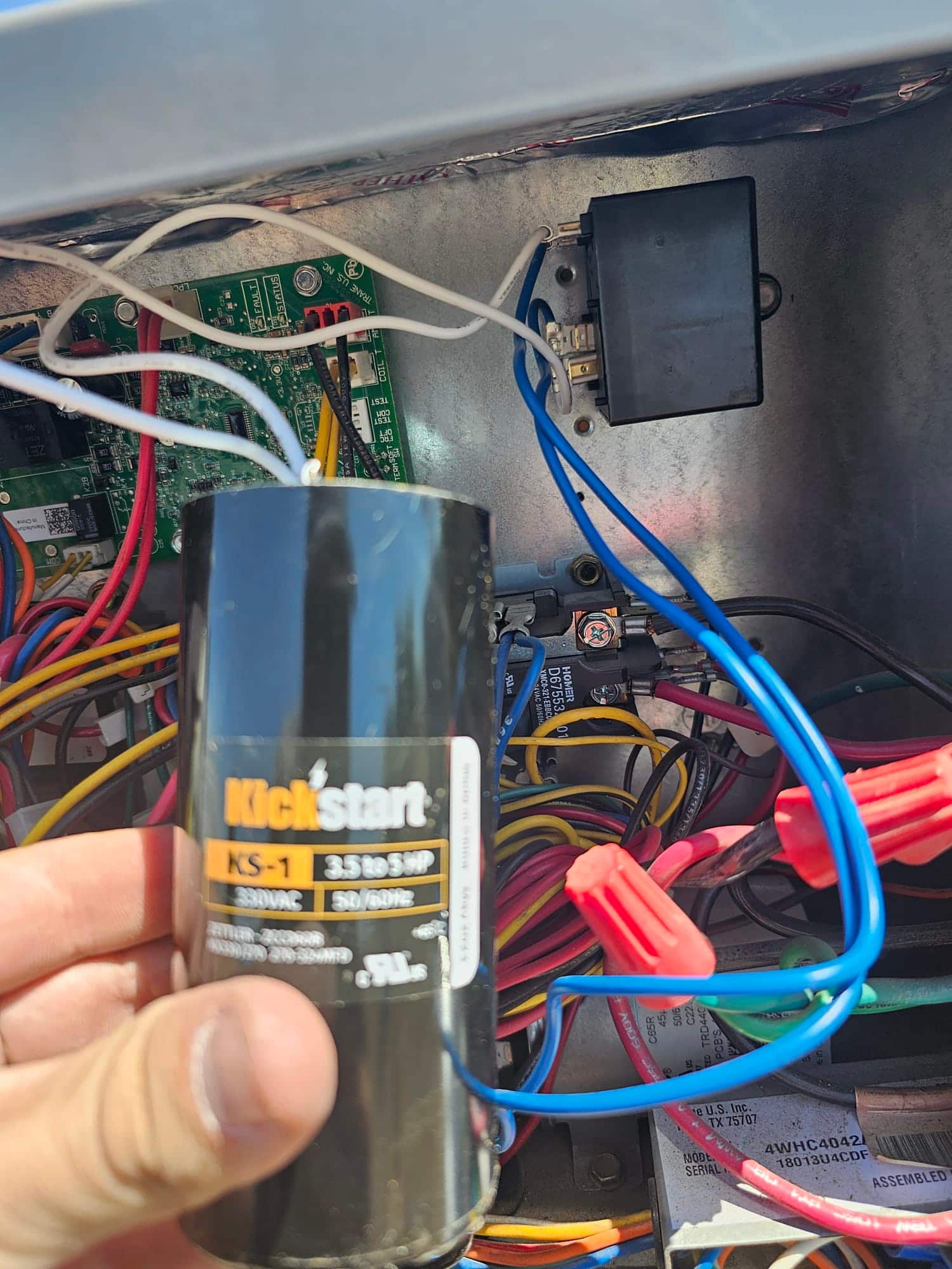 Technician holds a KS-1 start assist capacitor while wiring a relay into the outdoor condenser control compartment.