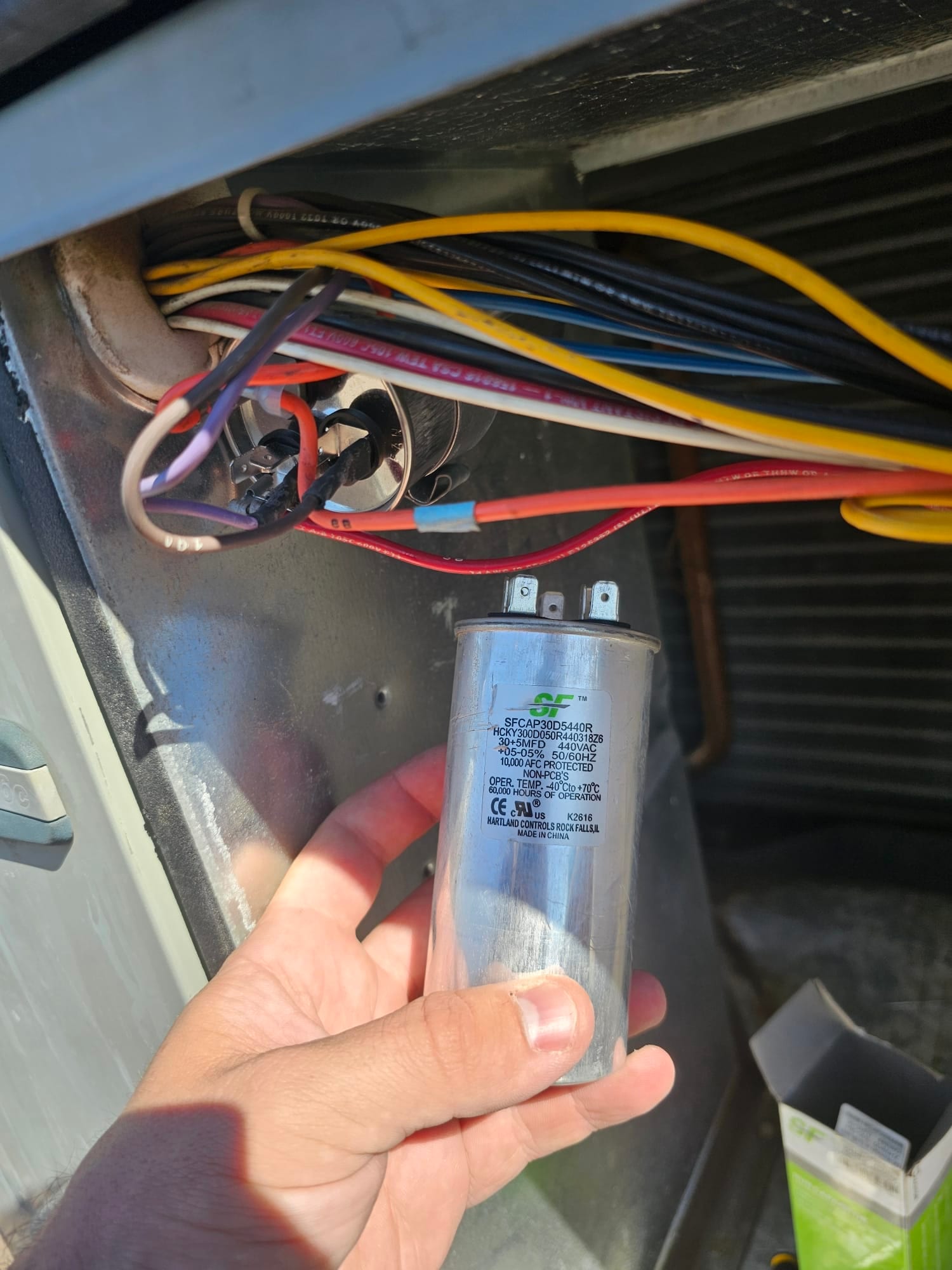 Technician holds a metal run capacitor near the condenser’s wiring compartment, showing connected wires and the evaporator coil in the background.