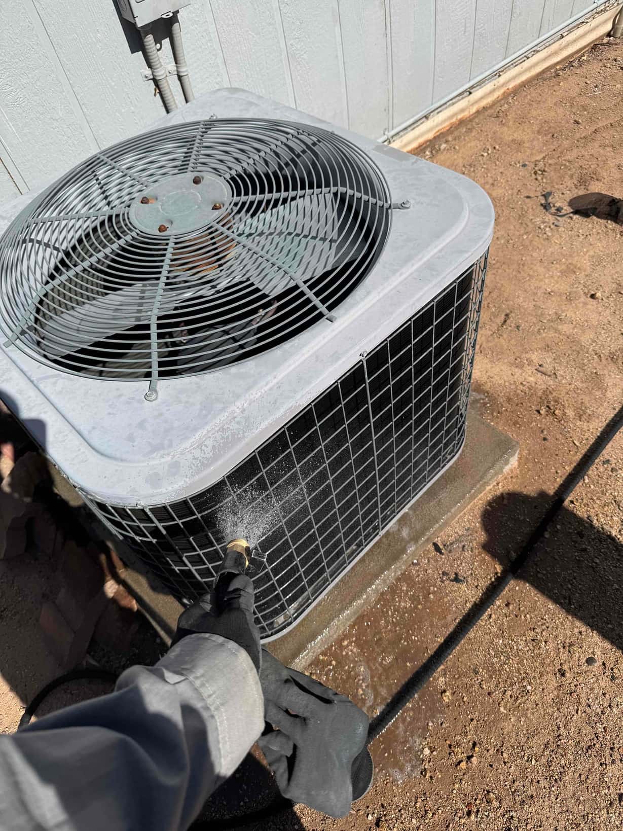 Technician sprays water to clean debris from the outdoor condenser coil and fan housing on a concrete pad.