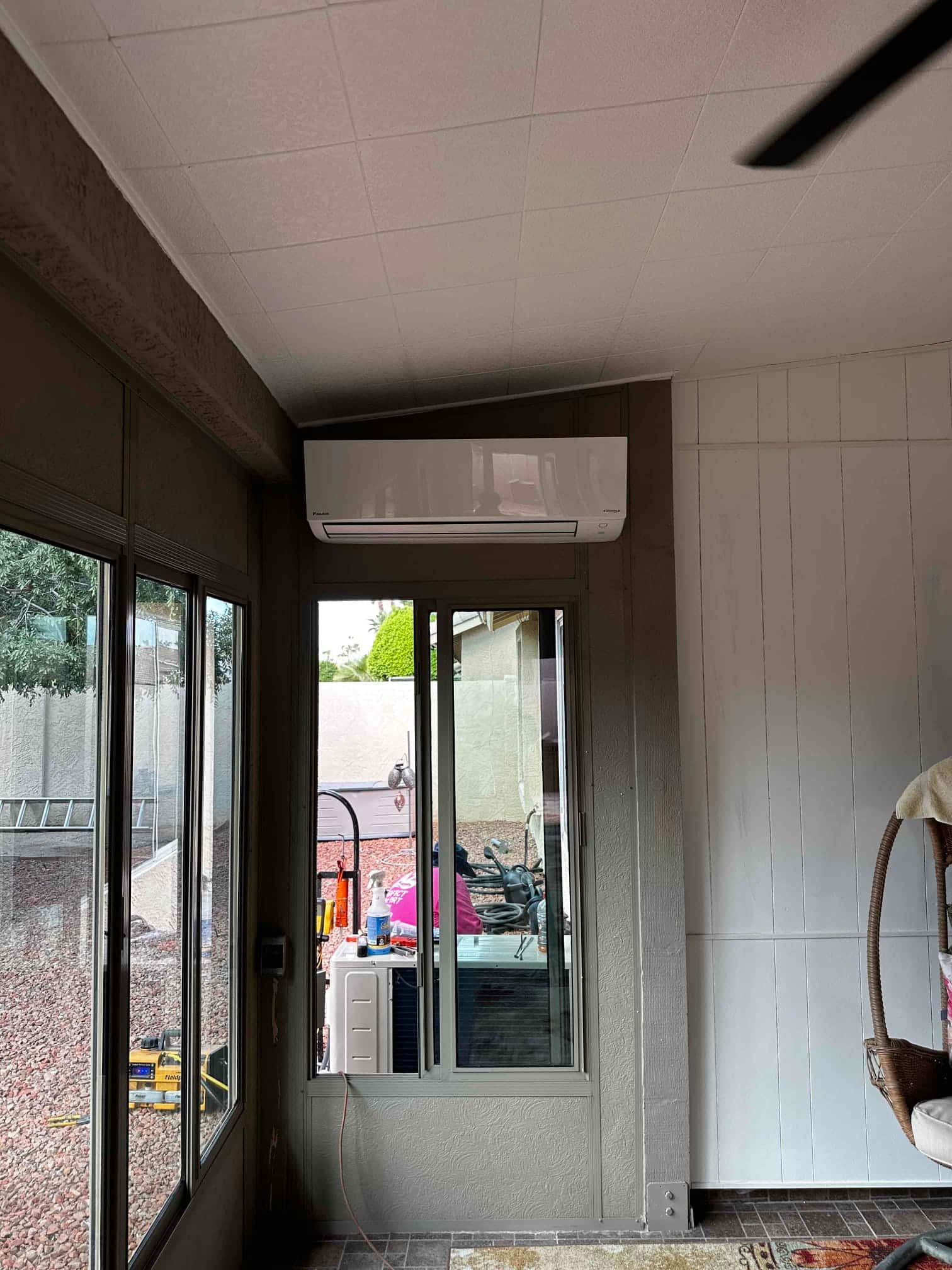 Wall-mounted mini-split indoor unit installed above a window in a finished room, with outdoor condenser visible through the glass.