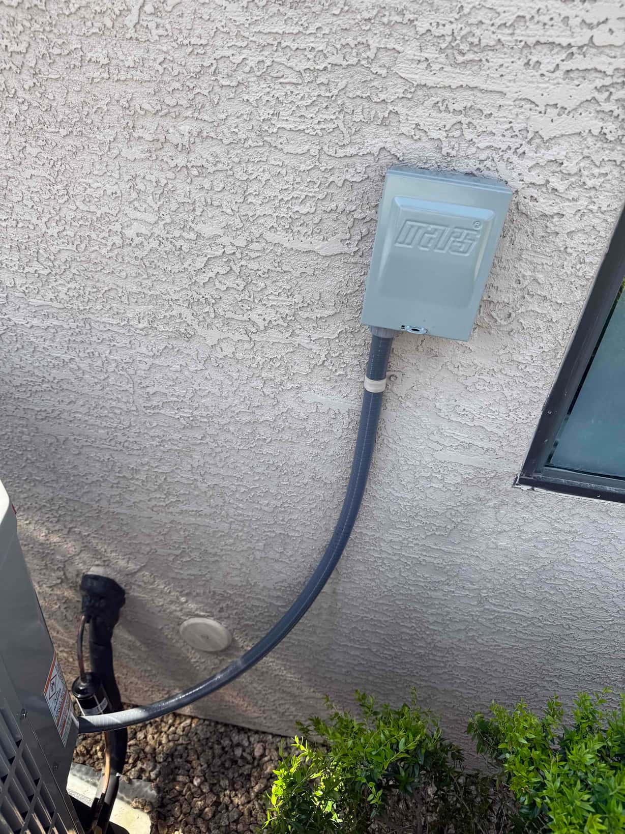 Weatherproof electrical disconnect with flexible conduit installed beside the outdoor condenser on the exterior wall.