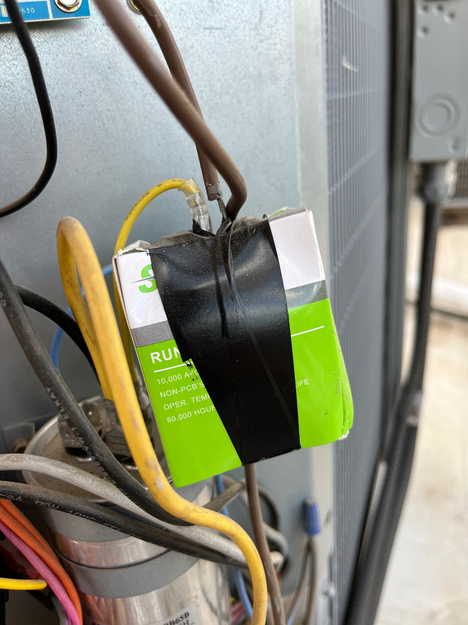 Boxed run capacitor taped in place, with yellow and brown conductors connected, alongside a cylindrical capacitor and wiring.
