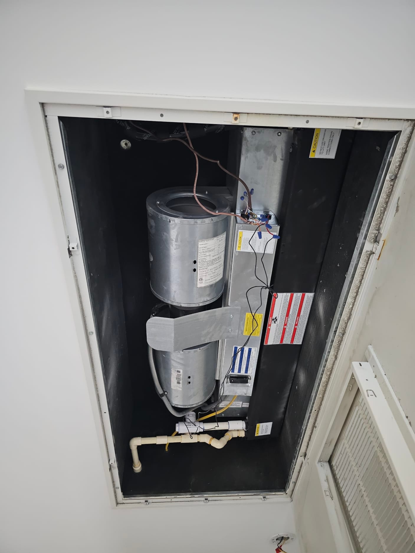 Ceiling-mounted air handler compartment open, showing blower housing, control wiring, and condensate drain piping.