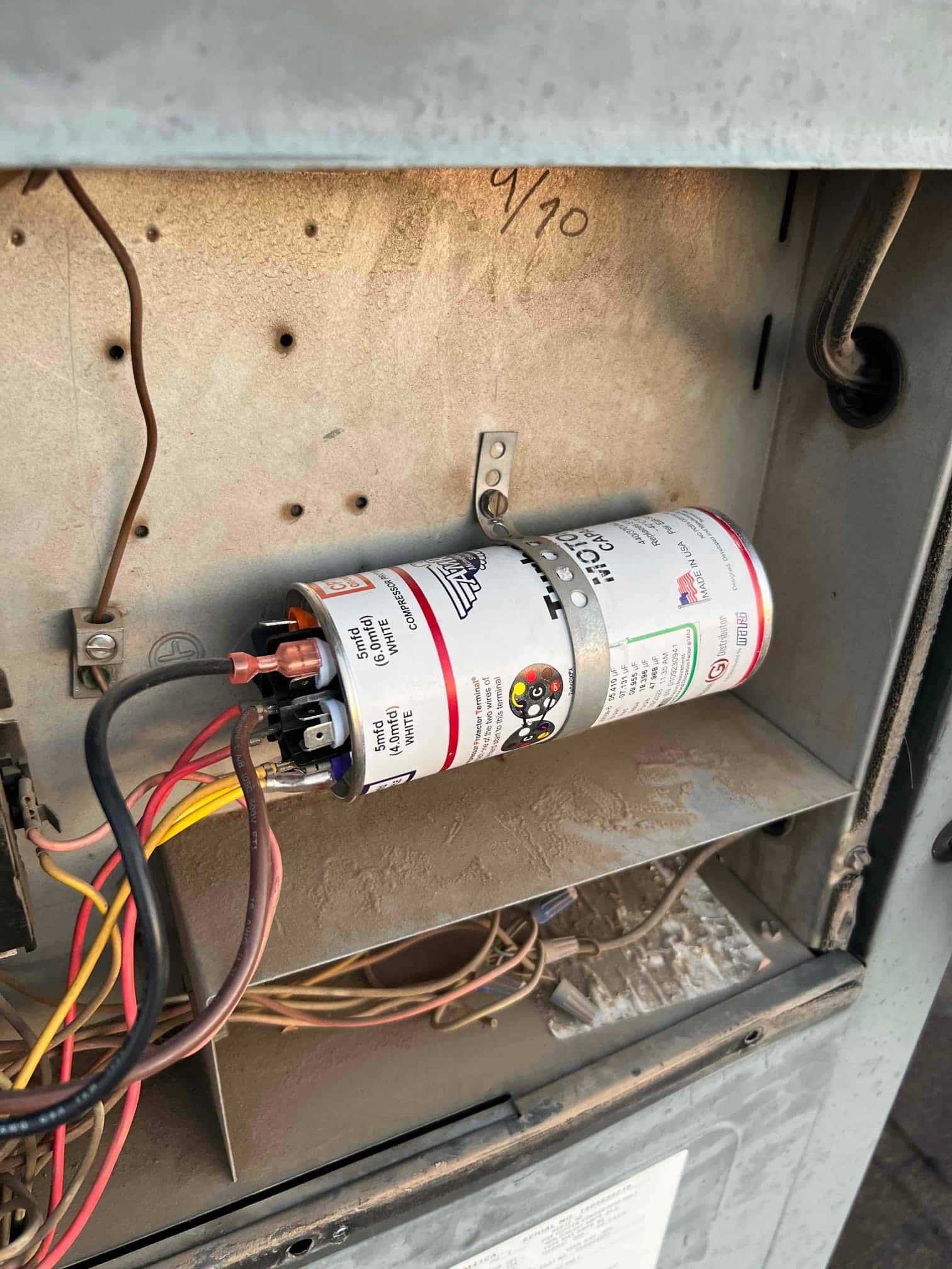 Cylindrical capacitor secured with a metal strap inside the unit, with multiple wires connected to its terminals. Dust present on interior surfaces.