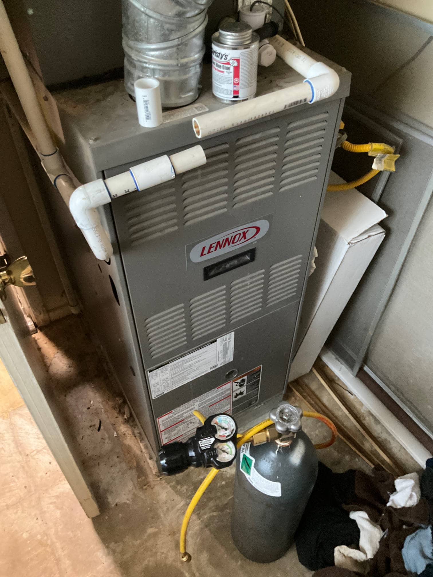 Furnace cabinet with PVC vent and condensate piping; gas cylinder with regulator and pressure gauges sits beside the unit with yellow hoses visible.