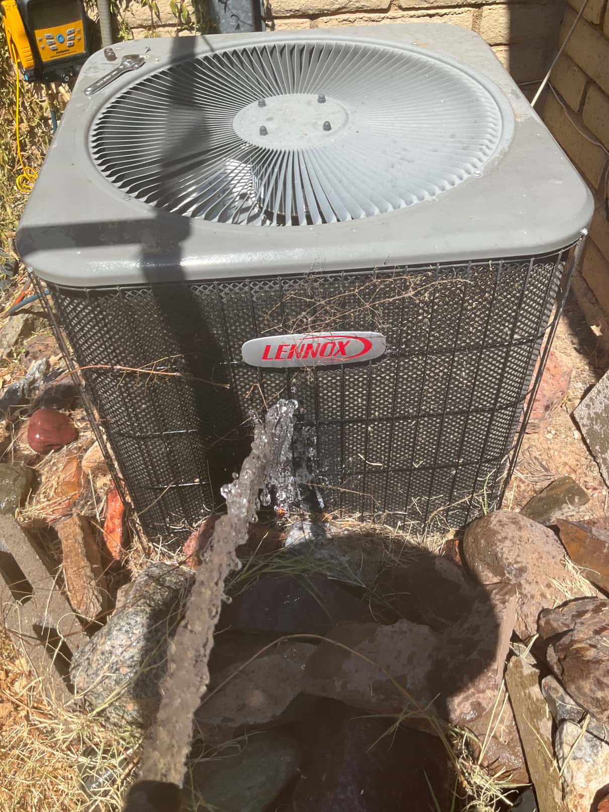 A garden hose sprays water across the outdoor condenser coil and grille.