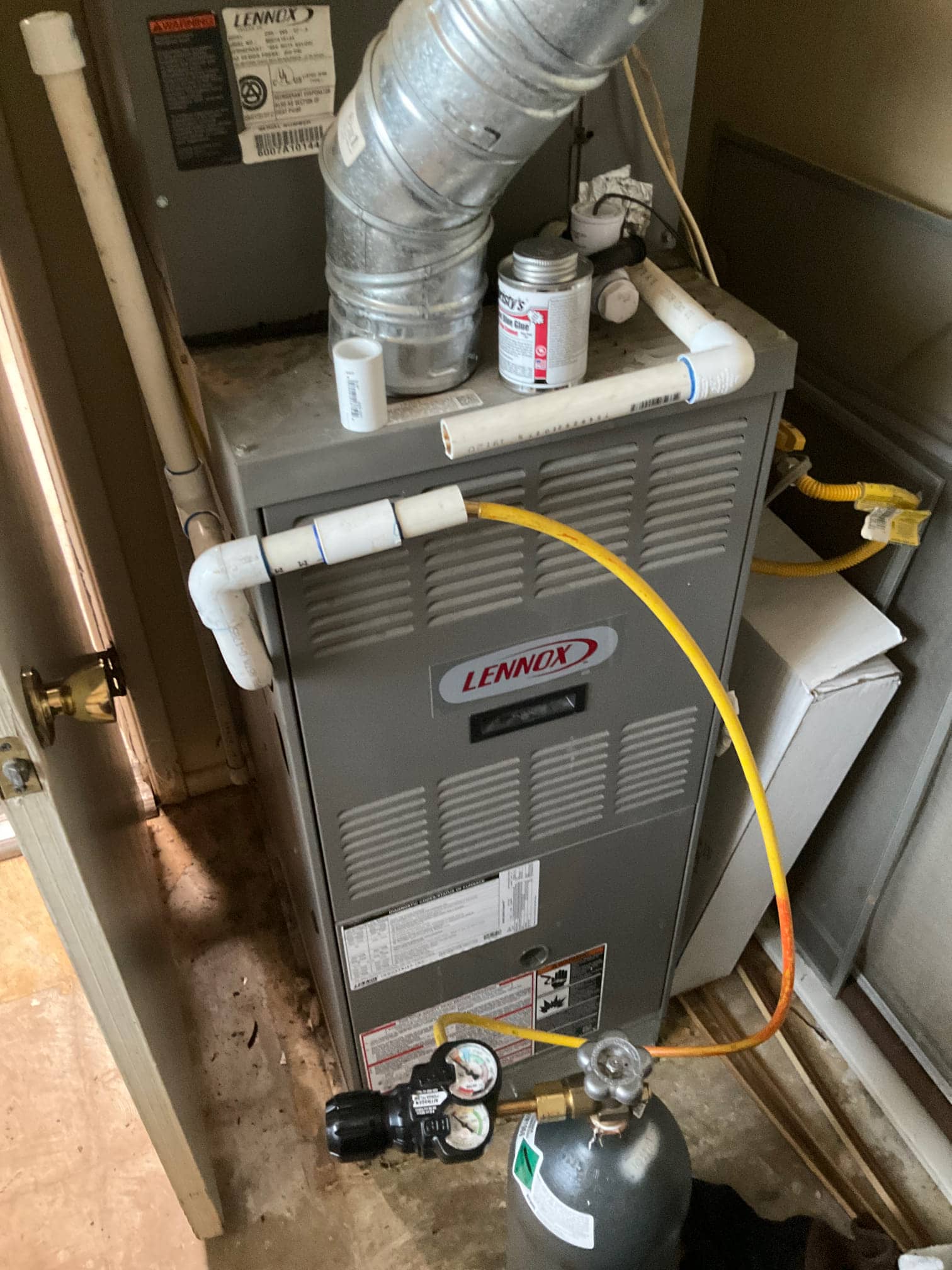 Indoor furnace cabinet with metal vent and PVC drain piping, plus a cylinder regulator with dual gauges and a connected yellow hose.