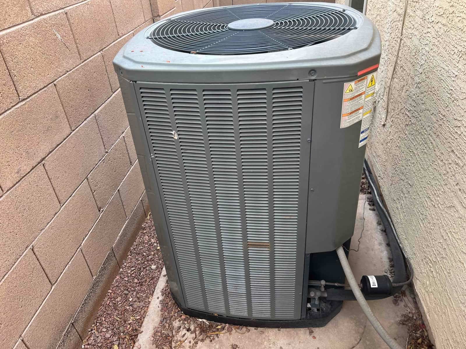 Outdoor air conditioner condenser beside the house, with refrigerant lines, service valves, and warning labels visible on the cabinet.