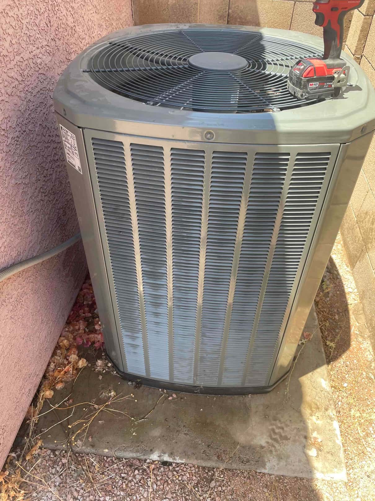 Outdoor air conditioner condenser beside stucco wall, with cordless drill and battery resting on top cover.