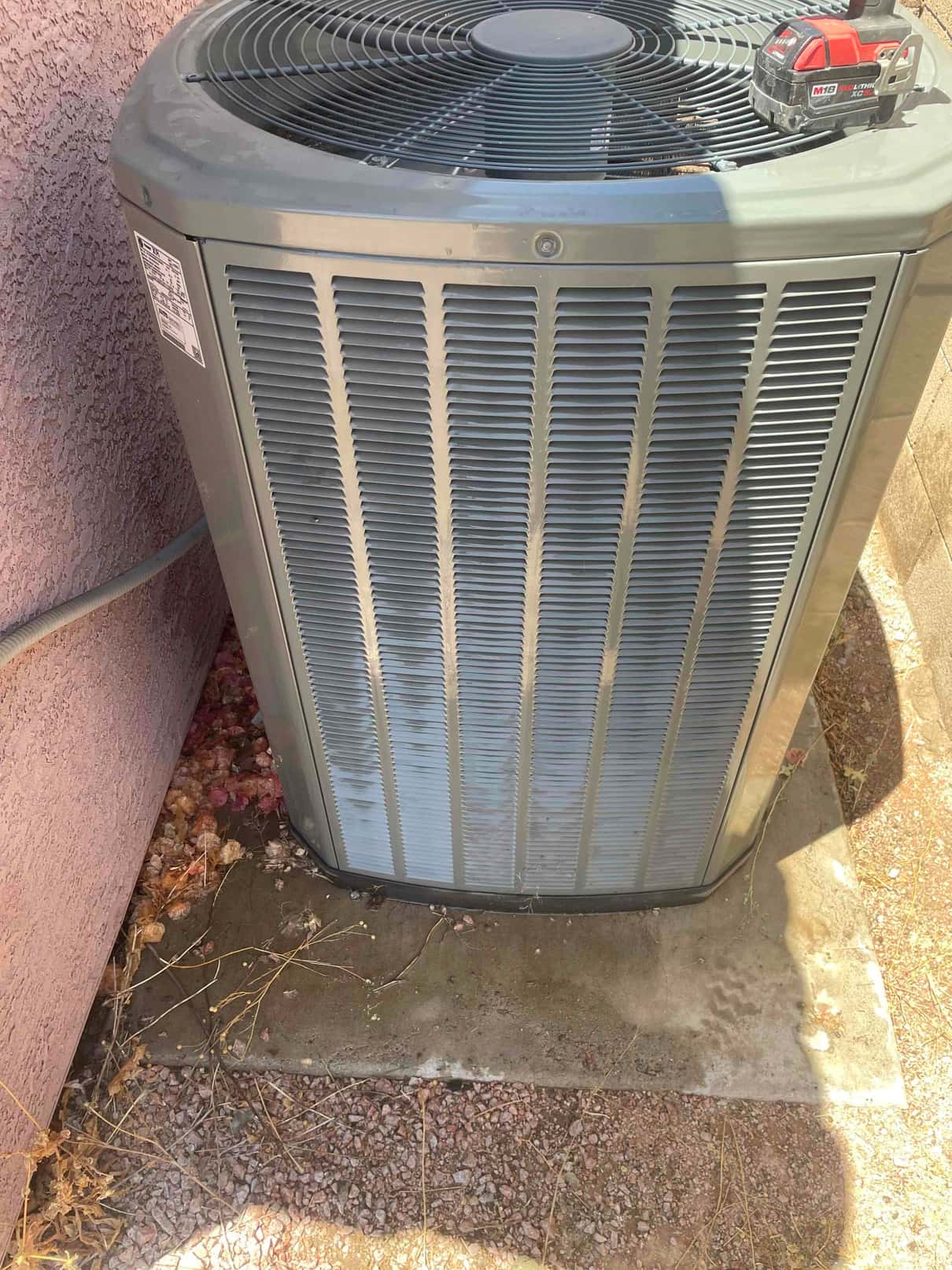 Outdoor air conditioner condenser unit beside stucco wall, fan grille on top, with leaves and debris around the base.