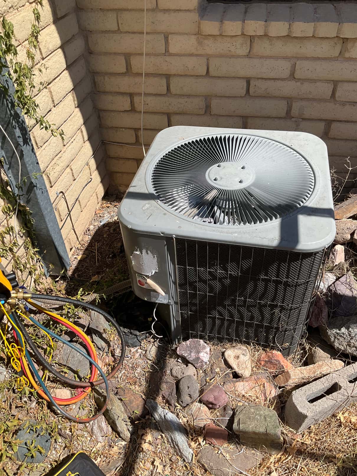 Outdoor air-conditioning condenser beside brick wall, top fan and coil visible, with refrigerant hoses positioned to the left.
