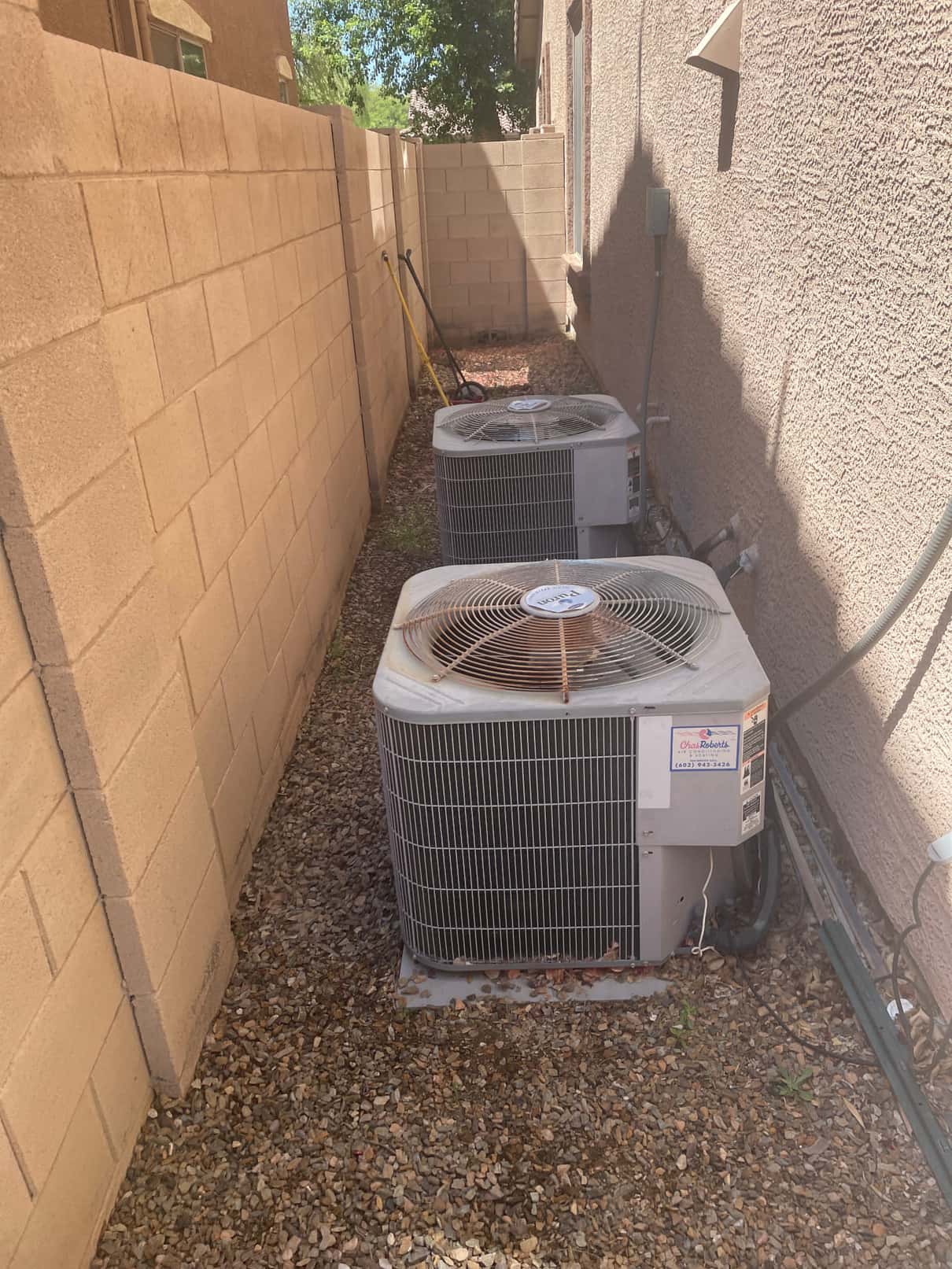 Two outdoor air conditioning condenser units along the house wall, with electrical disconnects and refrigerant lines visible; fan tops show leaves and surface rust.
