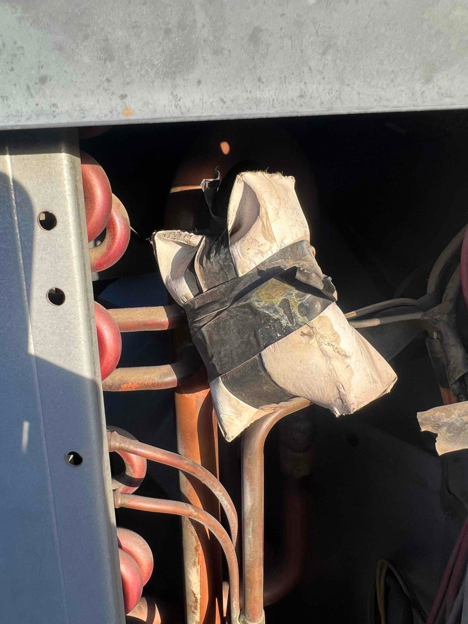 Outdoor condenser cabinet showing copper refrigerant lines and deteriorated, taped foam insulation with torn sections.
