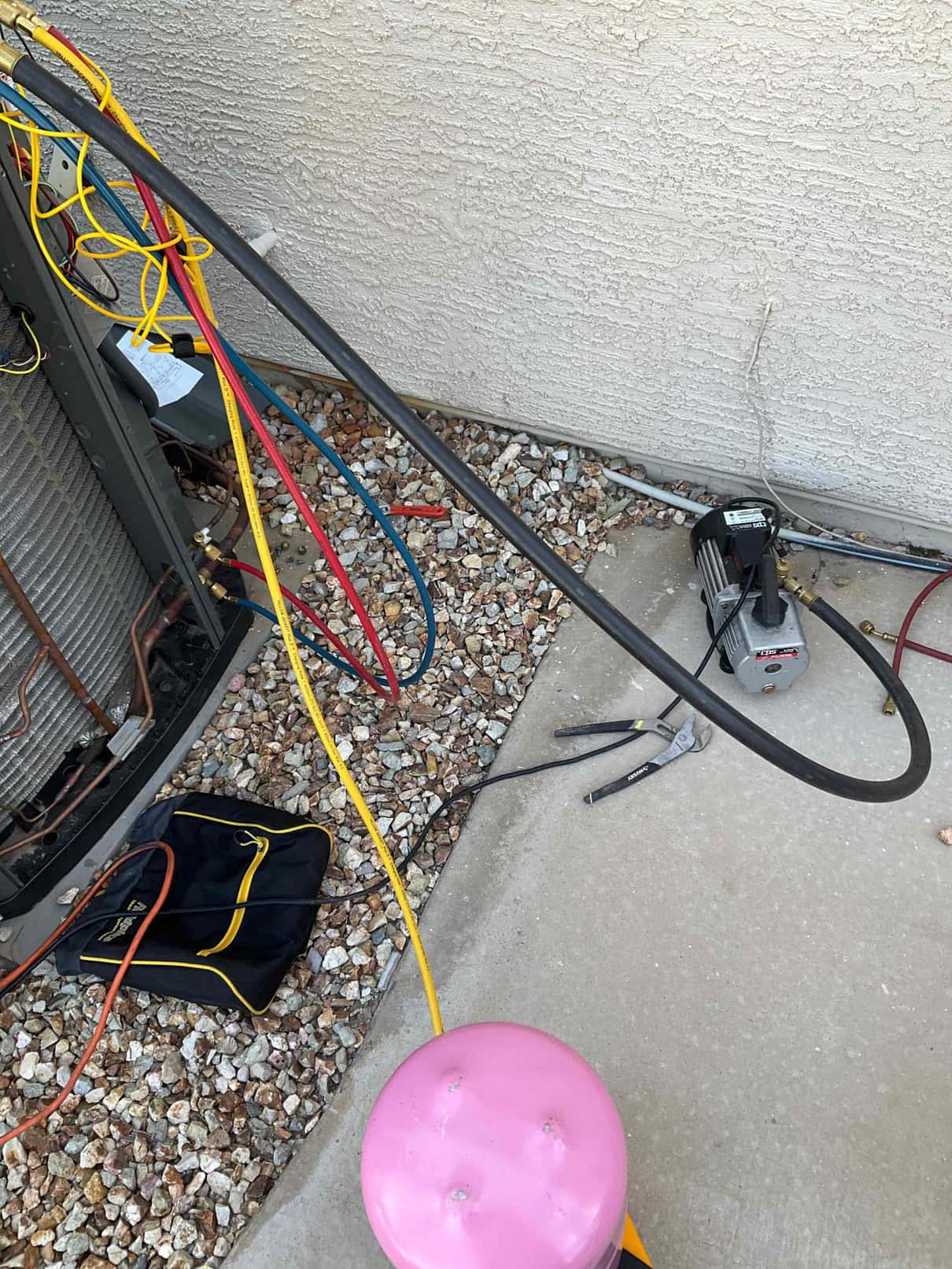 Outdoor condenser with service hoses connected; vacuum pump and pink refrigerant cylinder on the ground beside the unit.