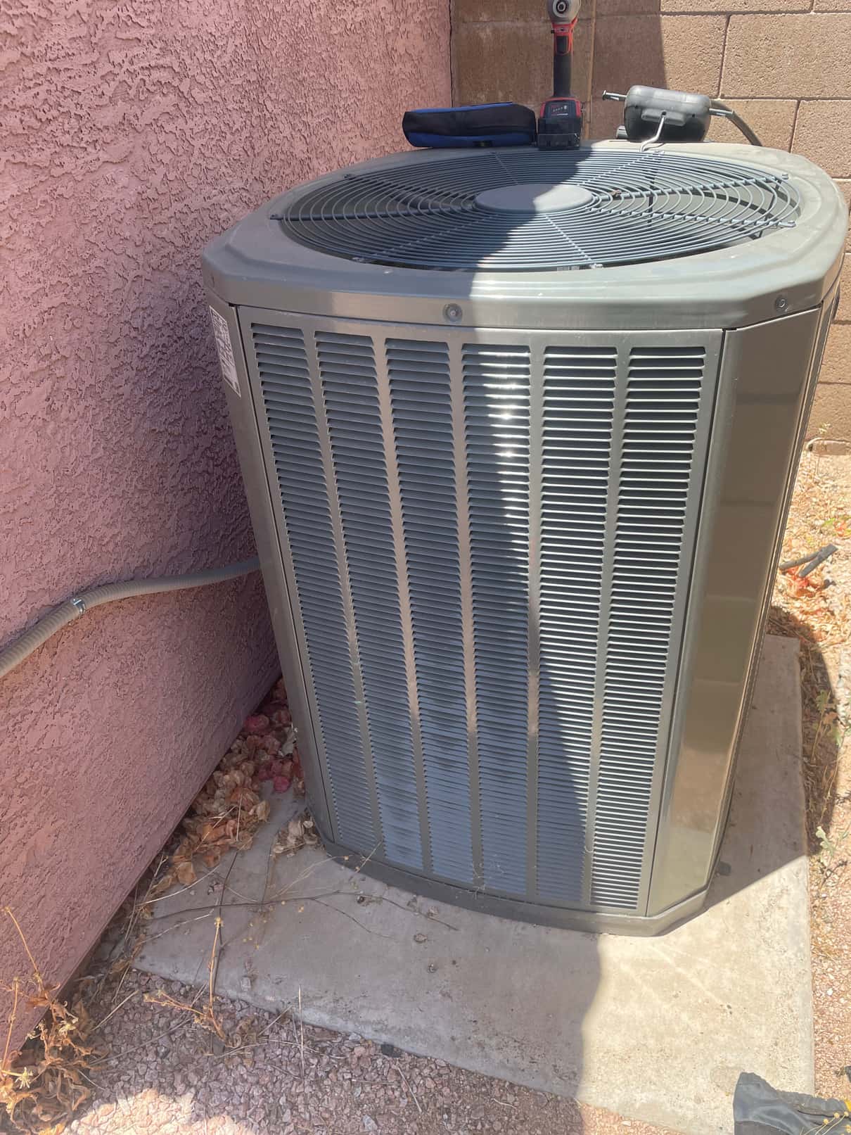 Outdoor condenser unit on a concrete pad beside a stucco wall, with tools resting on the fan grille.
