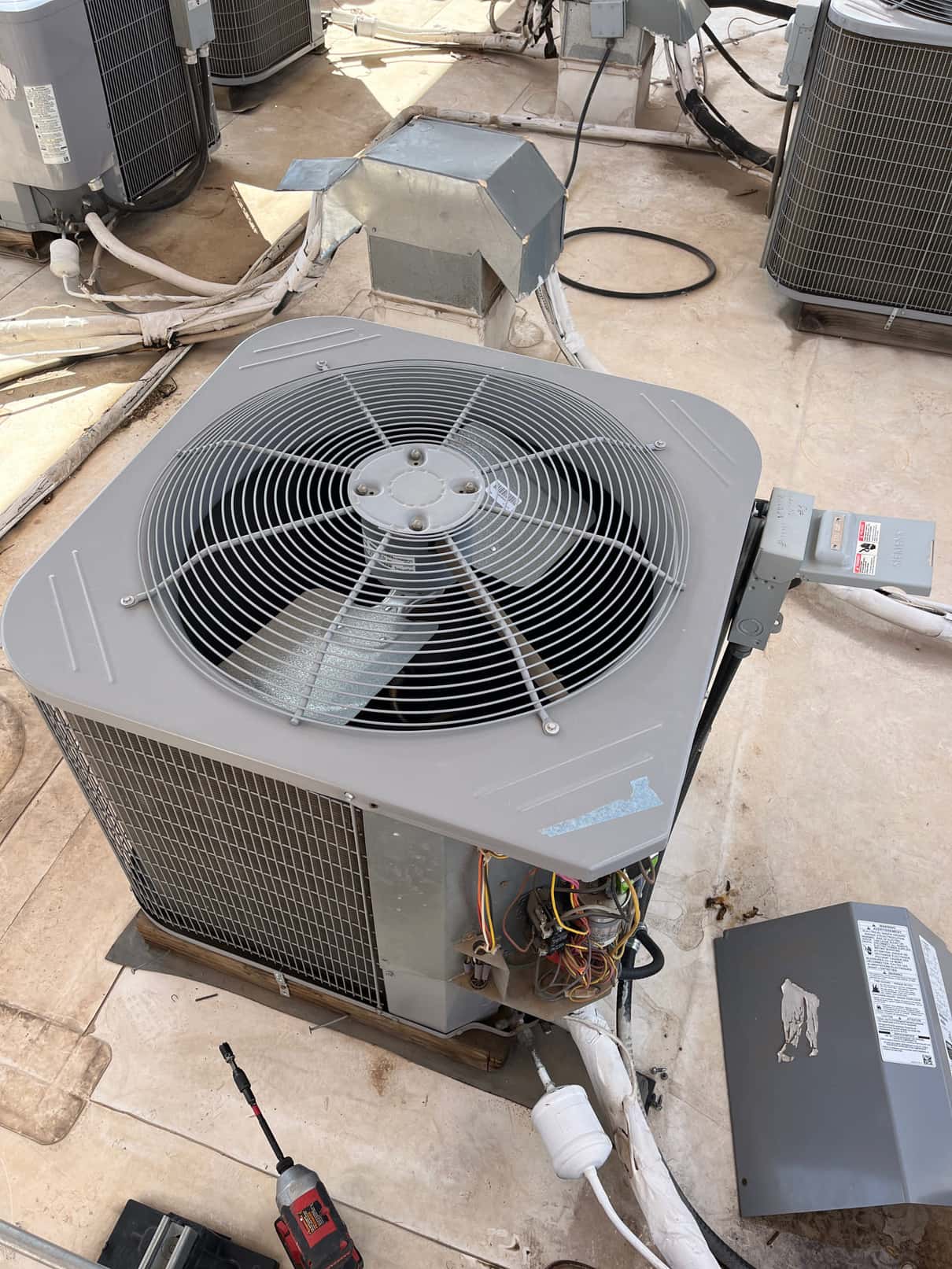 Outdoor condenser unit on rooftop with access panel removed, showing internal wiring and components; adjacent units and disconnect enclosure visible.