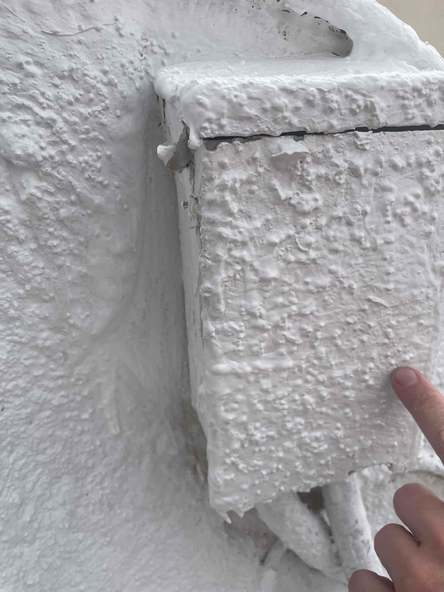 Outdoor electrical box and conduits on a stucco wall, heavily coated with white paint, technician’s finger pointing at the cover.