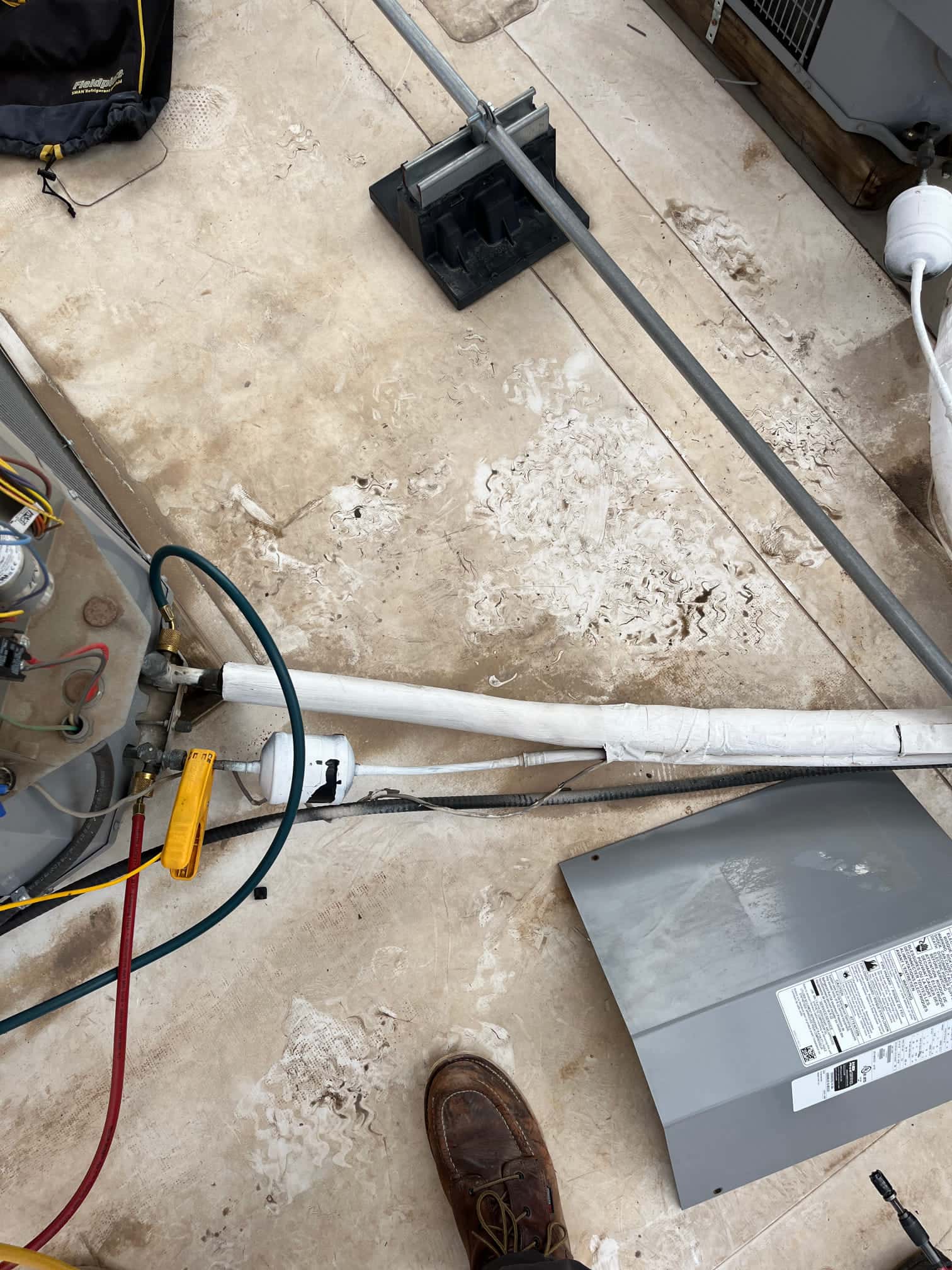 Outdoor HVAC work area showing insulated refrigerant lines, service hoses connected, exposed wiring, and the equipment access panel removed on the ground.