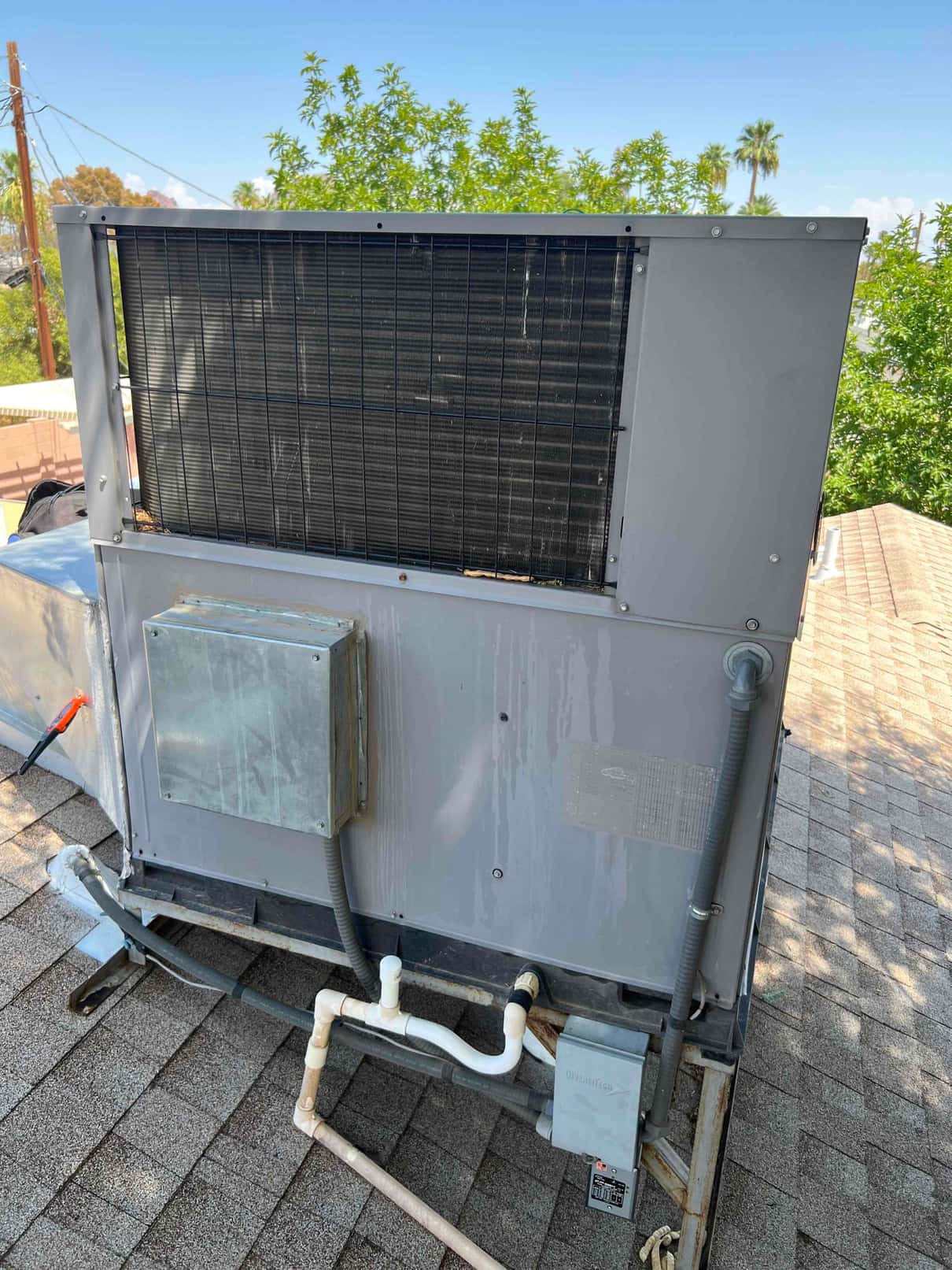 Rooftop packaged HVAC unit showing condenser coil, electrical junction box, flexible conduits, service disconnect, and PVC condensate drain piping on the shingle roof.