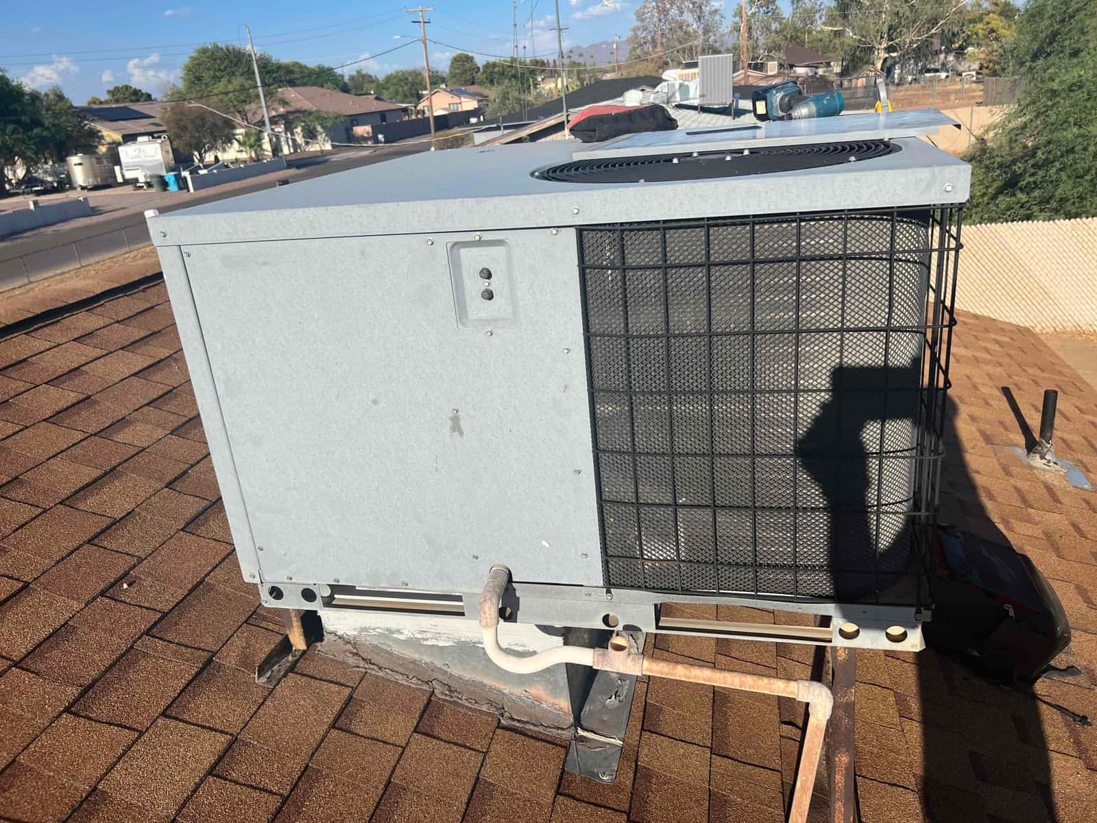 Rooftop packaged HVAC unit on shingles, condenser fan at top with protective grille and visible copper refrigerant piping connections.