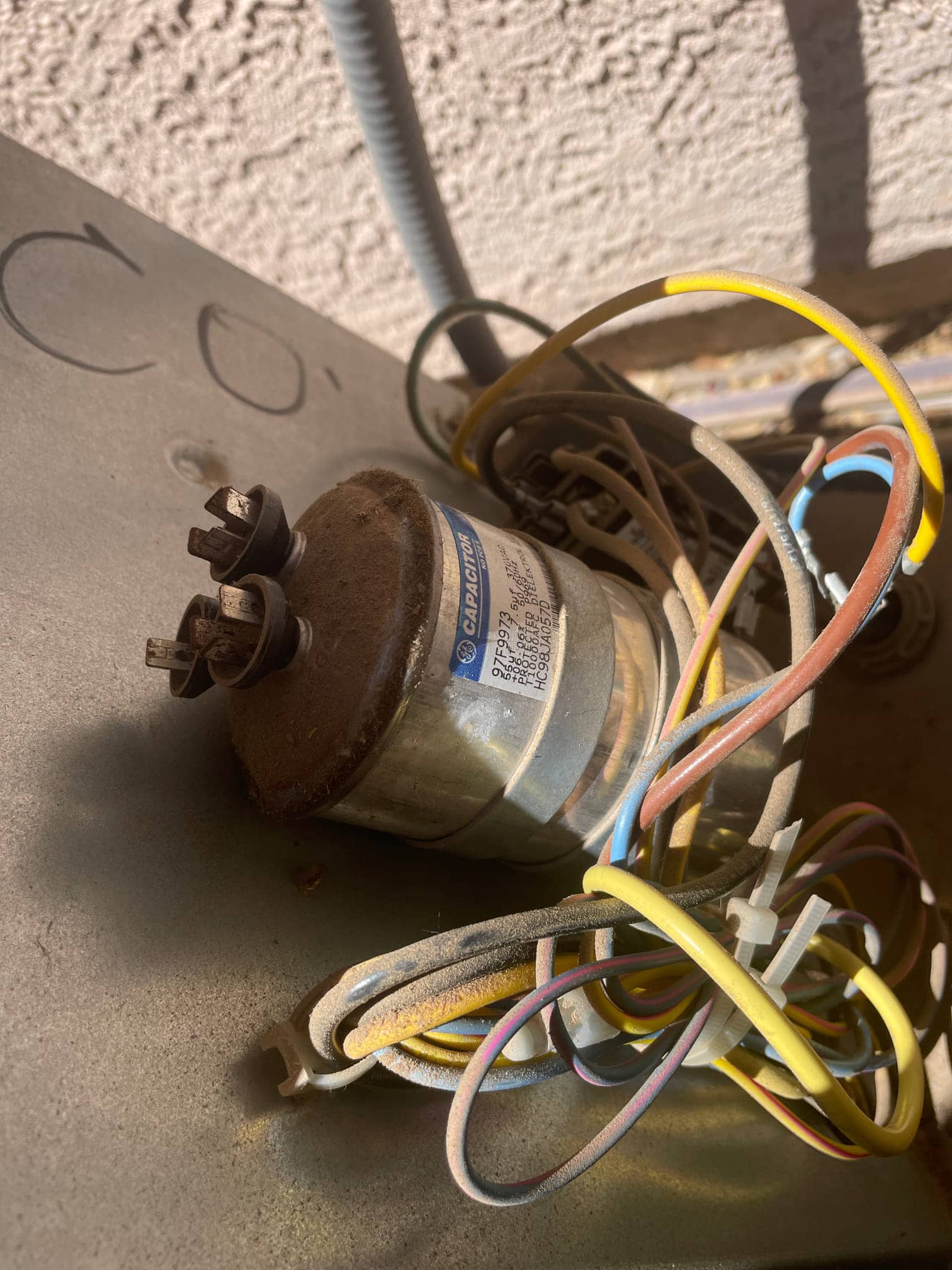 Round capacitor mounted inside outdoor unit compartment with corroded terminals and dusty, color-coded wires tied nearby.