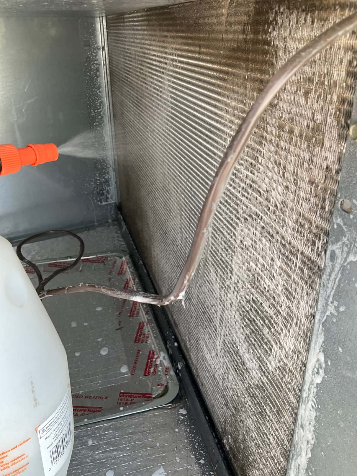 Spray nozzle applies foaming cleaner onto the evaporator coil inside the air handler, with drain pan visible below.