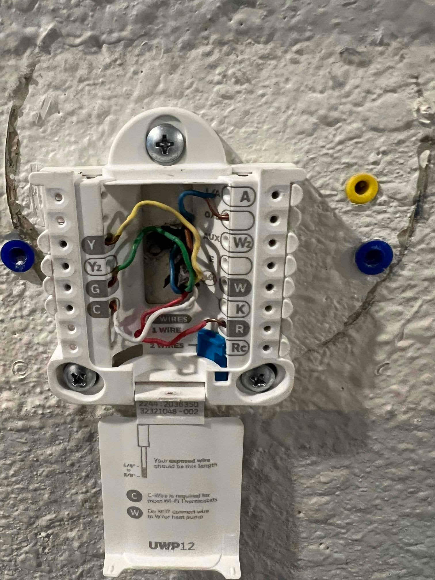 Thermostat mounting base on interior wall showing exposed multi-color control wires and labeled terminals including R, Rc, W, C, G, and Y.