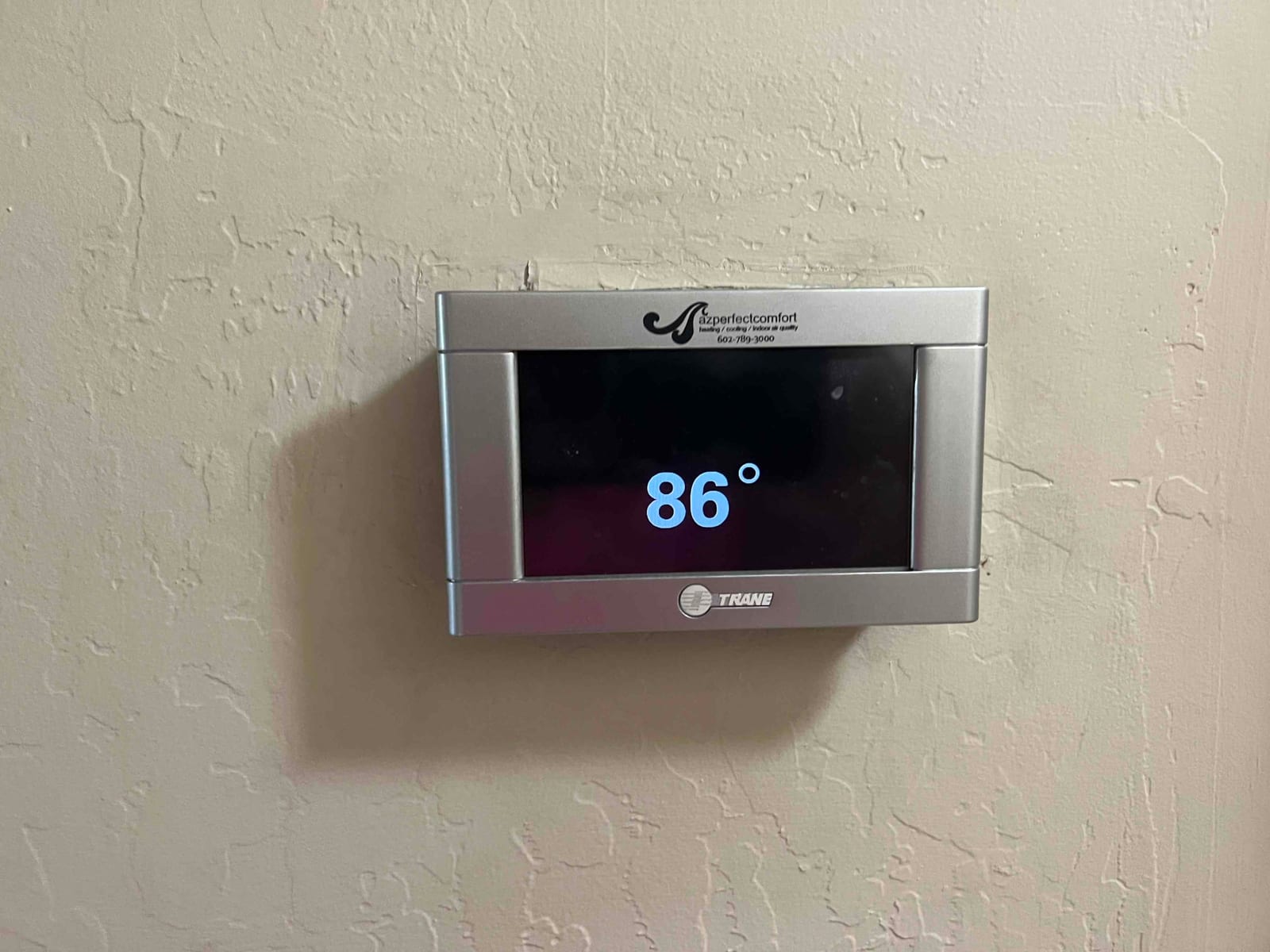 Wall-mounted thermostat touchscreen displays 86° in large numerals, mounted on textured interior wall.