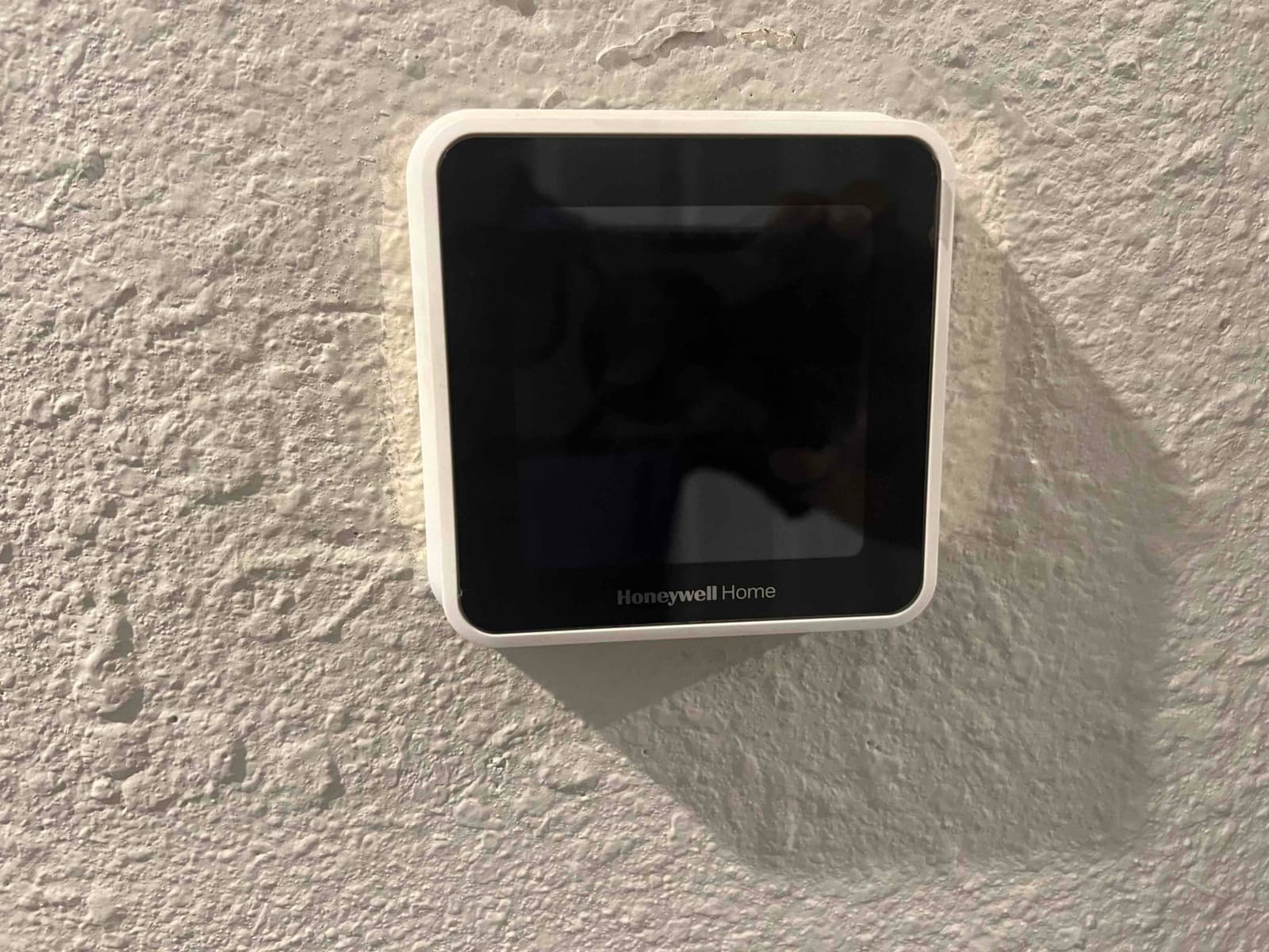 Wall-mounted touchscreen thermostat with dark display and white frame on a textured interior wall.
