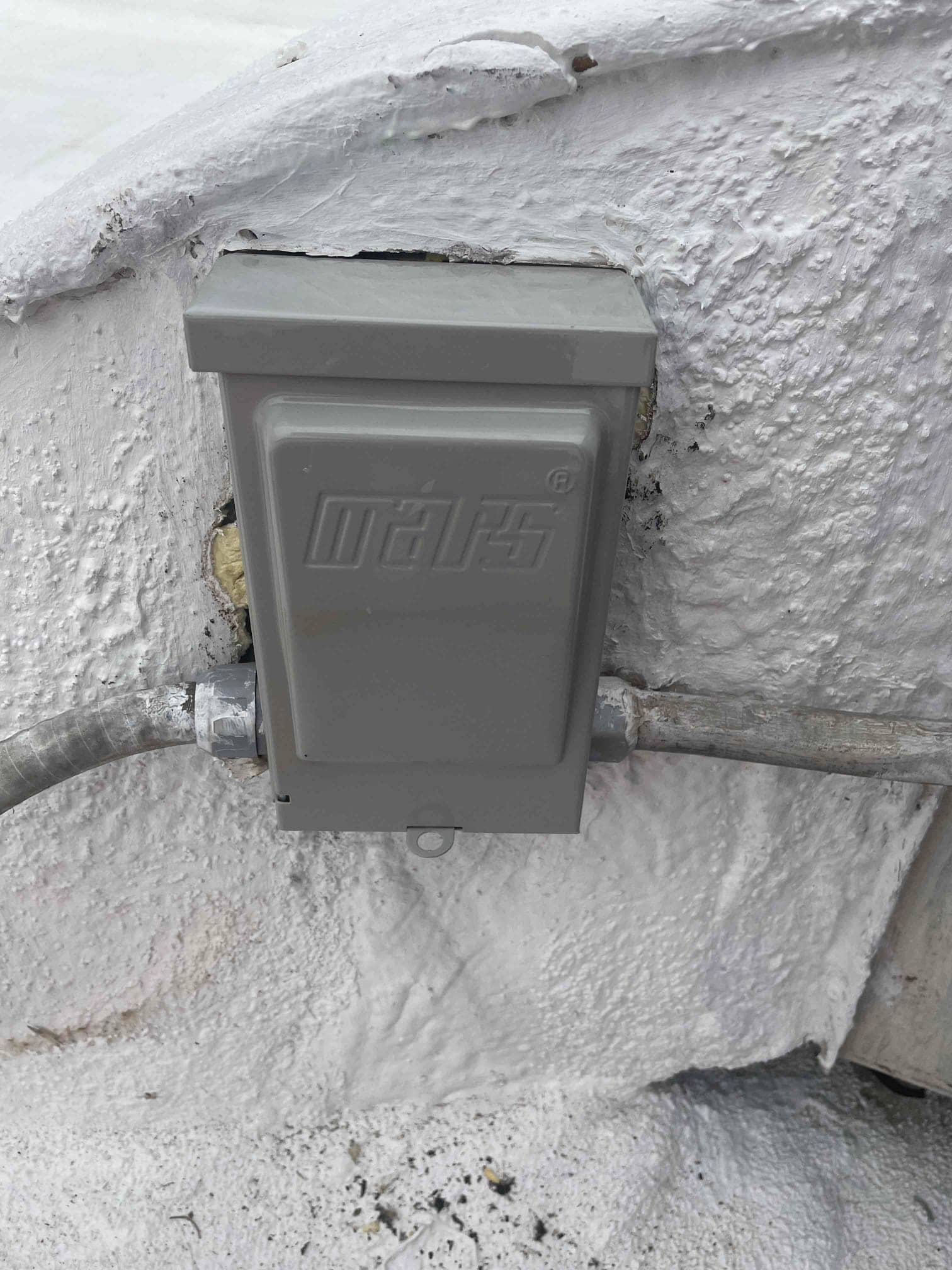 Weatherproof air-conditioner disconnect box mounted on exterior wall, with flexible metallic conduit entering both sides and sealed around penetration.