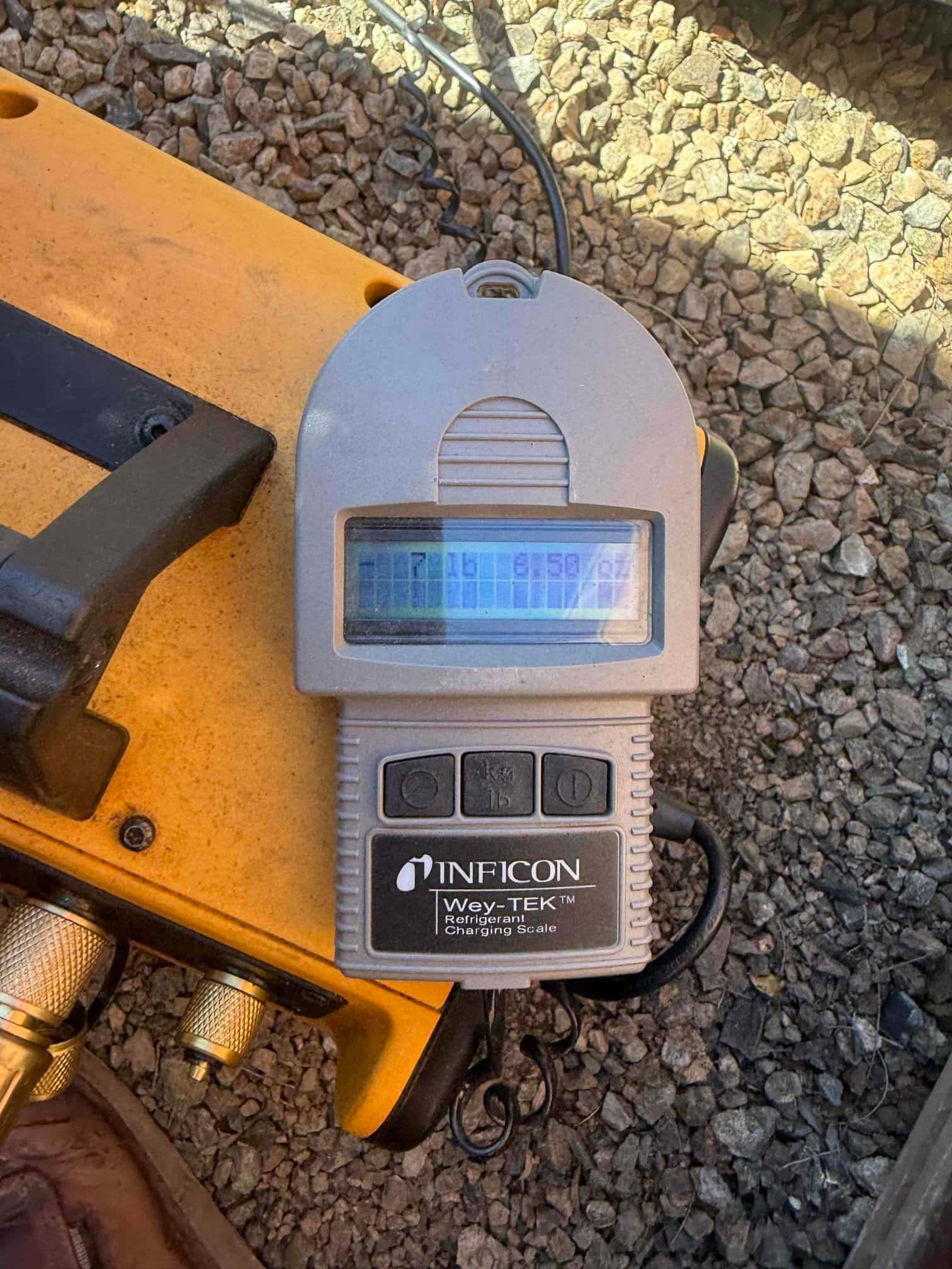 Handheld refrigerant scale display shown on the job site next to hoses and tools, used during system charging.