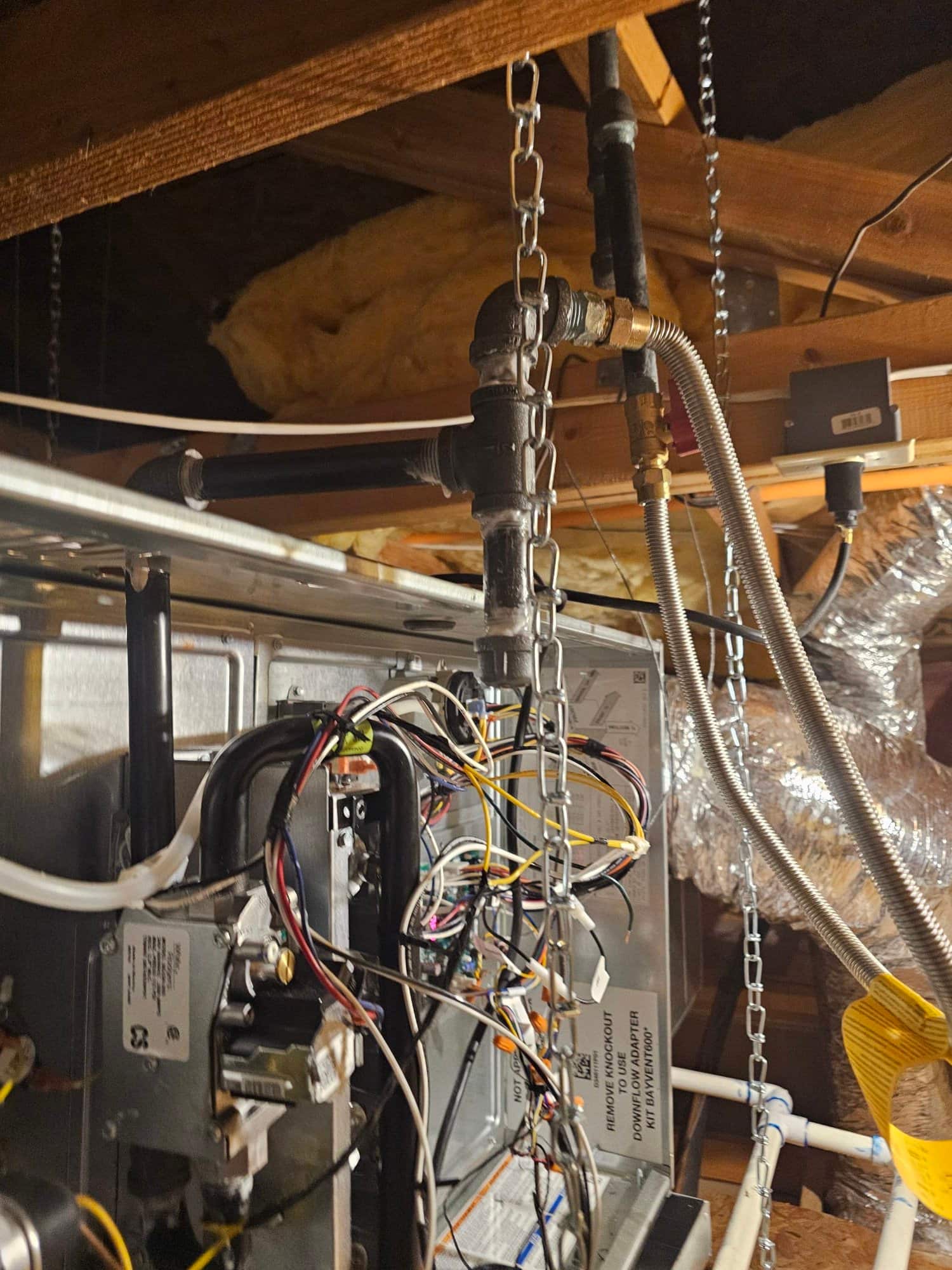 Attic air handler installation showing gas piping with flexible connector, drain piping, suspended support chains, and cabinet wiring exposed.