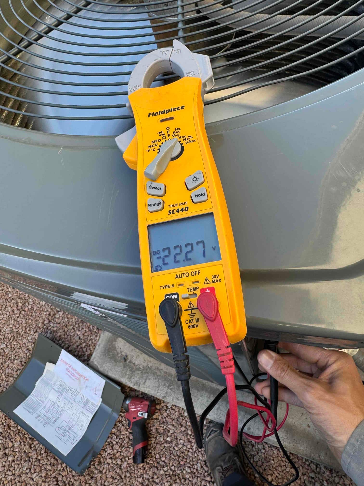 Clamp meter measures 222.7 V across two test leads at the outdoor condenser unit.